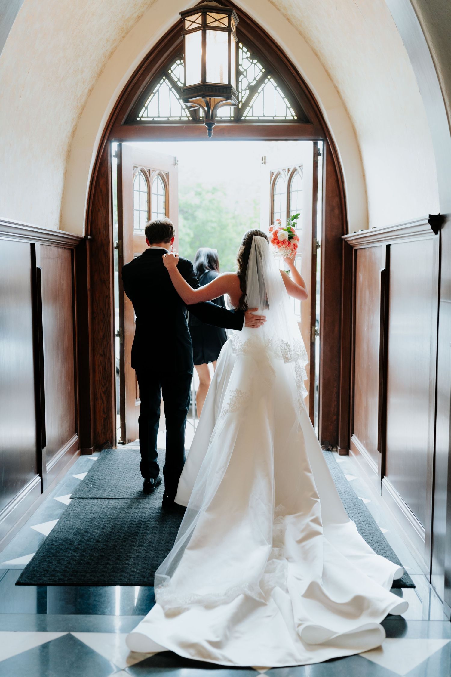 Bride in white gown with veil and groom in black suit walk toward church doors during wedding ceremony.