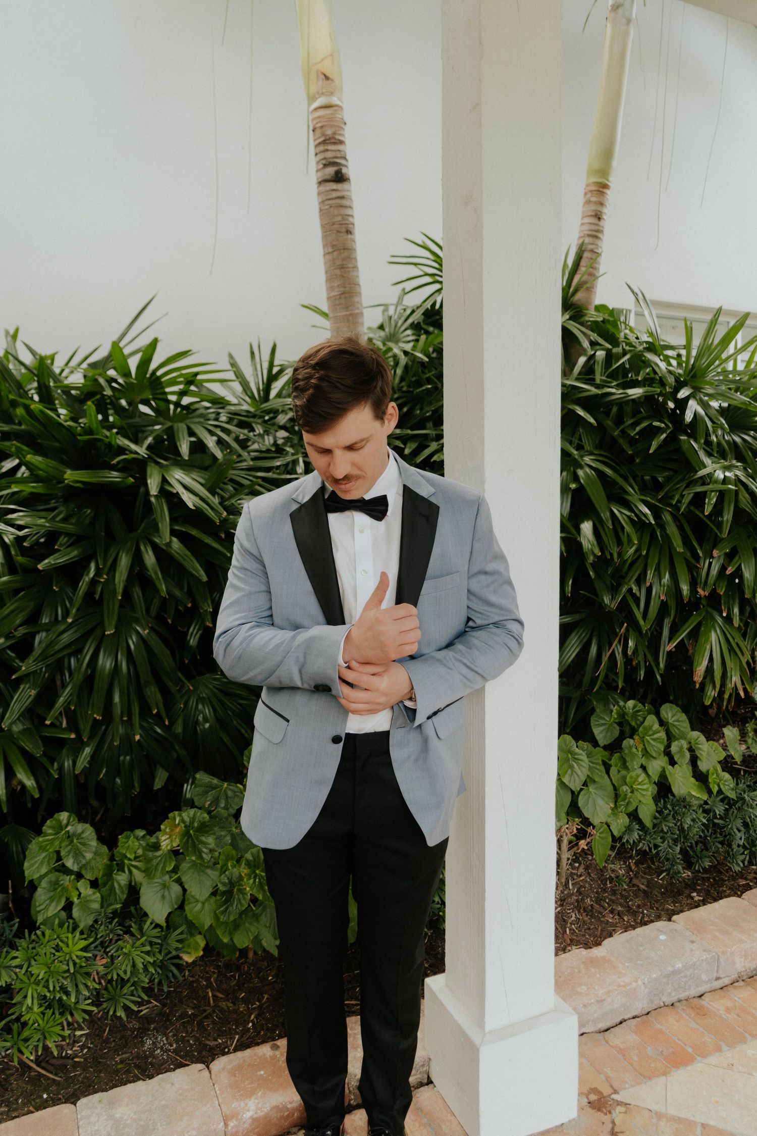 A groom in a light gray tuxedo jacket with black lapels and bow tie adjusts his cufflinks outdoors.