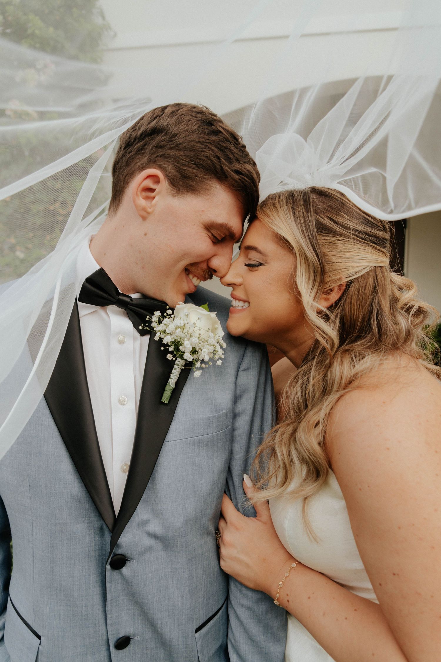 A smiling bride and groom touch foreheads under a flowing veil, groom in gray tuxedo with boutonniere, bride in white gown.