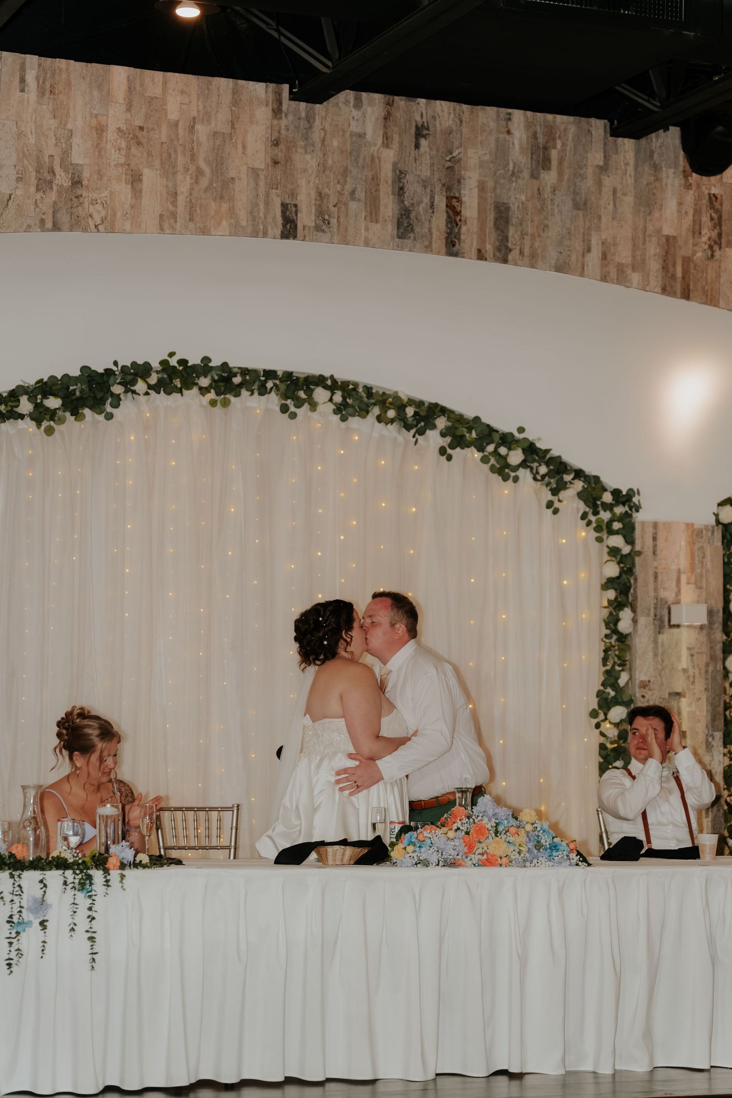 Bride and groom share a kiss at the sweetheart table beneath a floral arch with fairy lights at their wedding reception.