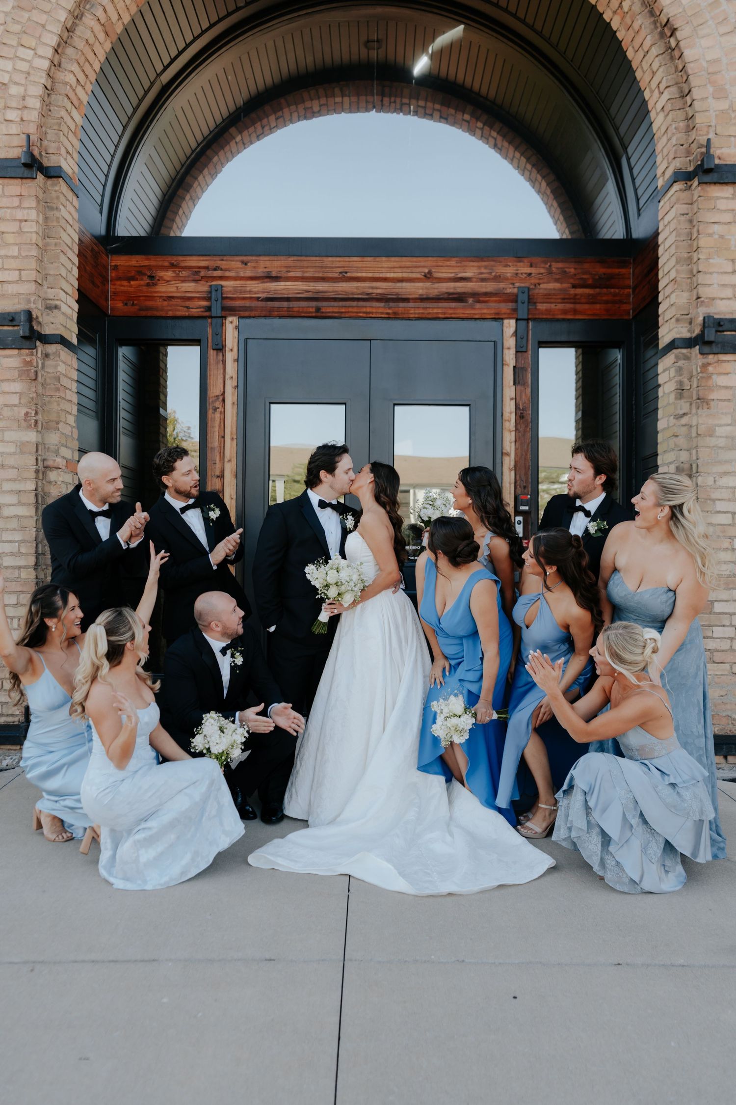 Wedding party poses outside brick venue, bride and groom kissing while bridesmaids in blue dresses and groomsmen in tuxedos celebrate.