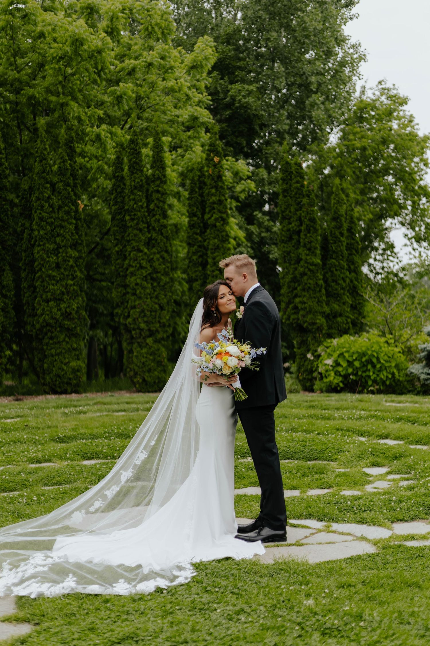 Bride and groom embrace in lush garden, bride wearing flowing white gown with long veil holding colorful bouquet.