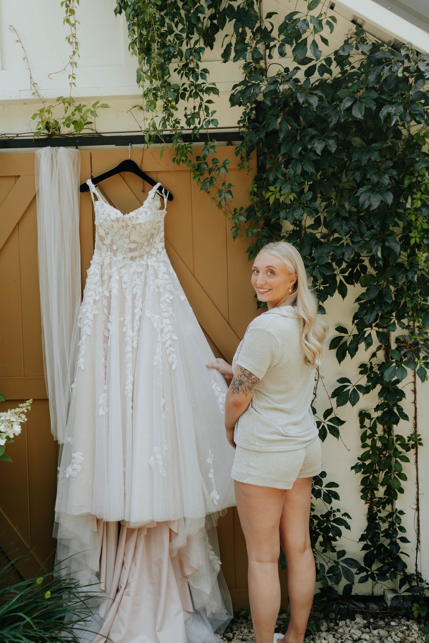 Bride admiring her floral lace wedding gown hanging outdoors against a yellow barn door with lush greenery.