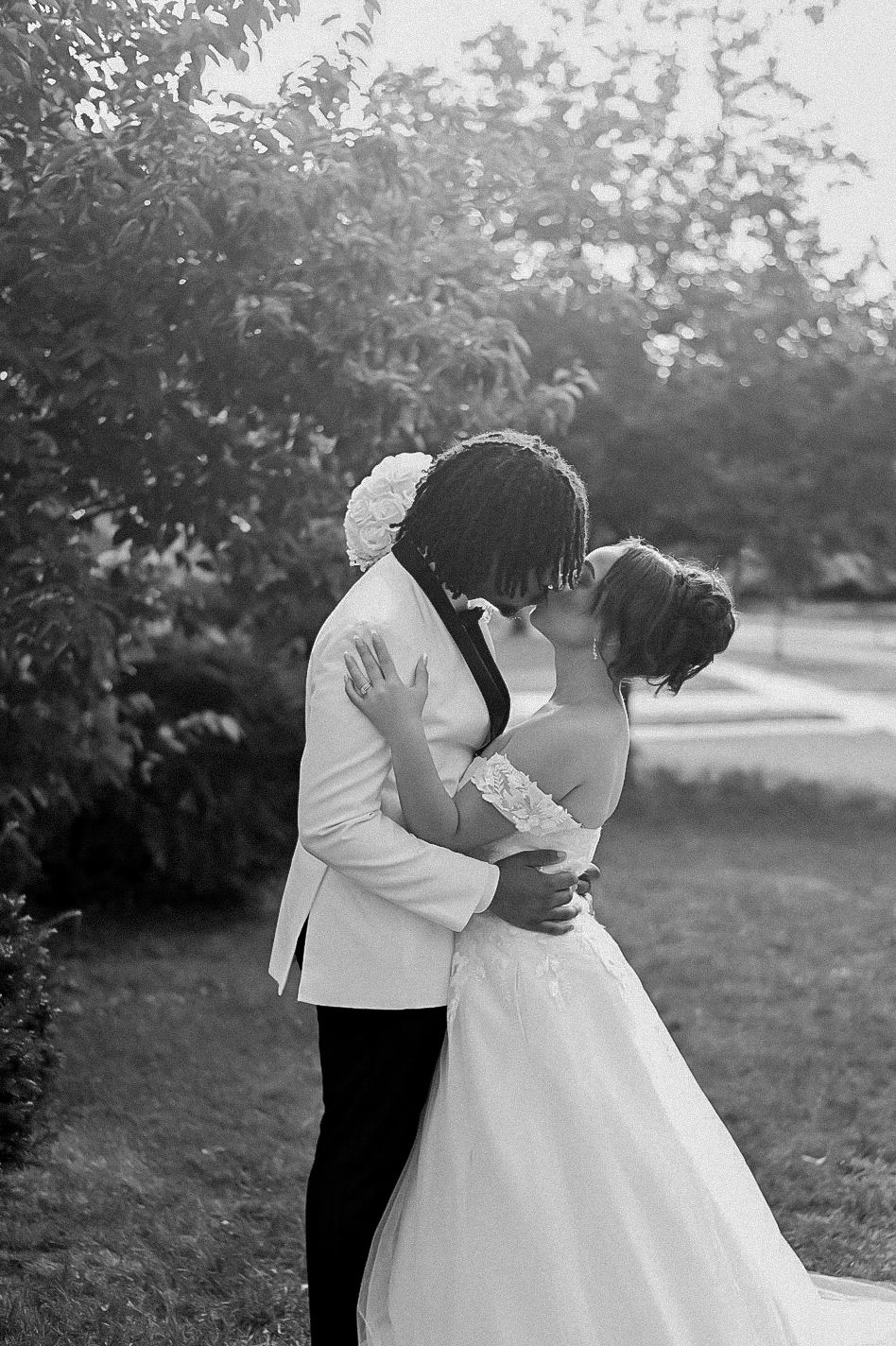 Black and white photo of a bride and groom sharing a romantic kiss outdoors, surrounded by lush greenery.