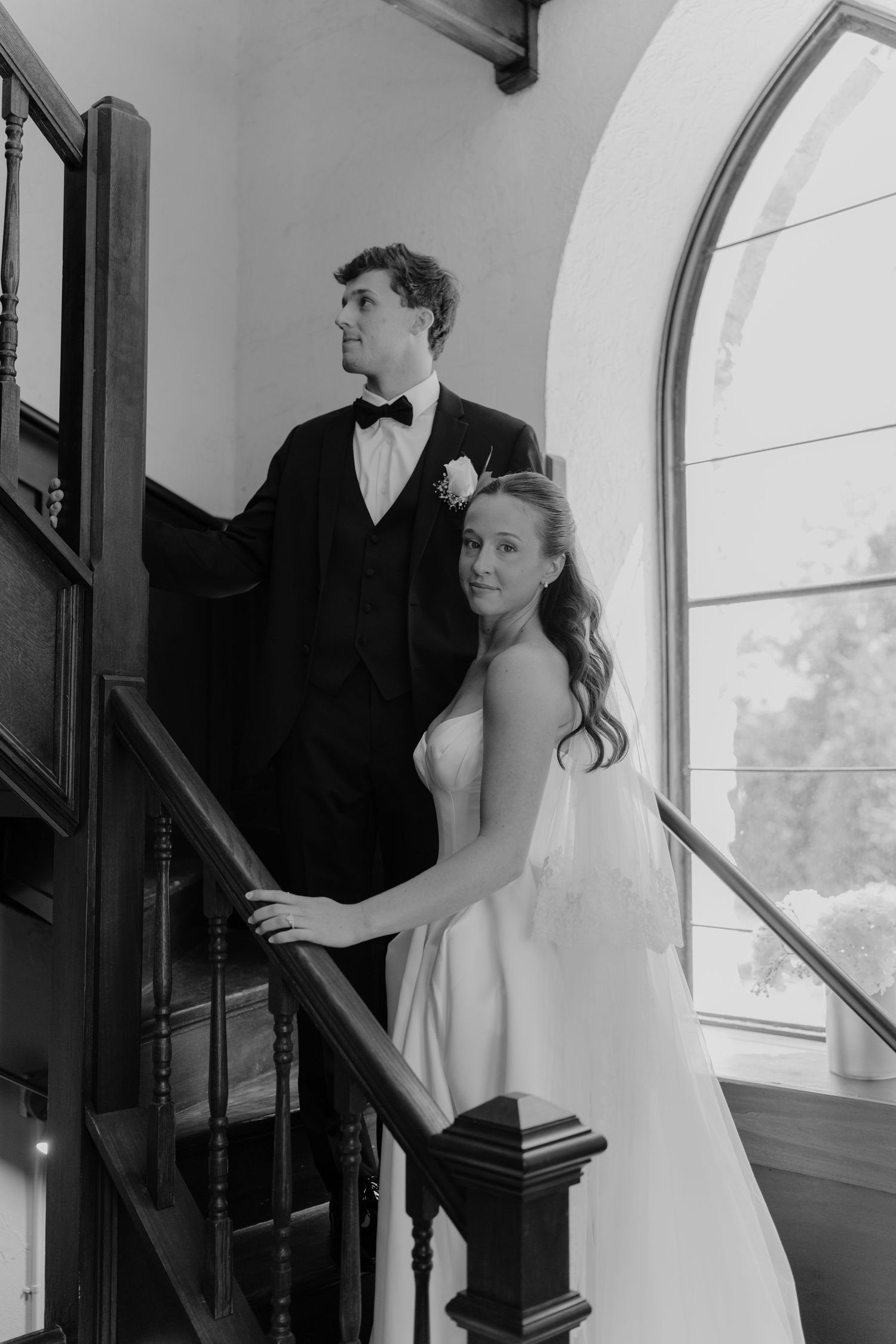 Black and white wedding photo of bride and groom posing elegantly on a staircase near an arched window.