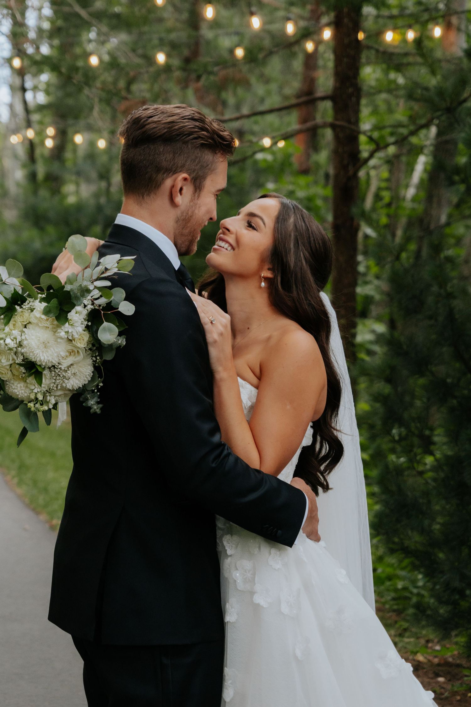 Bride and groom share a romantic embrace on a forest path, with string lights and lush greenery in the background.