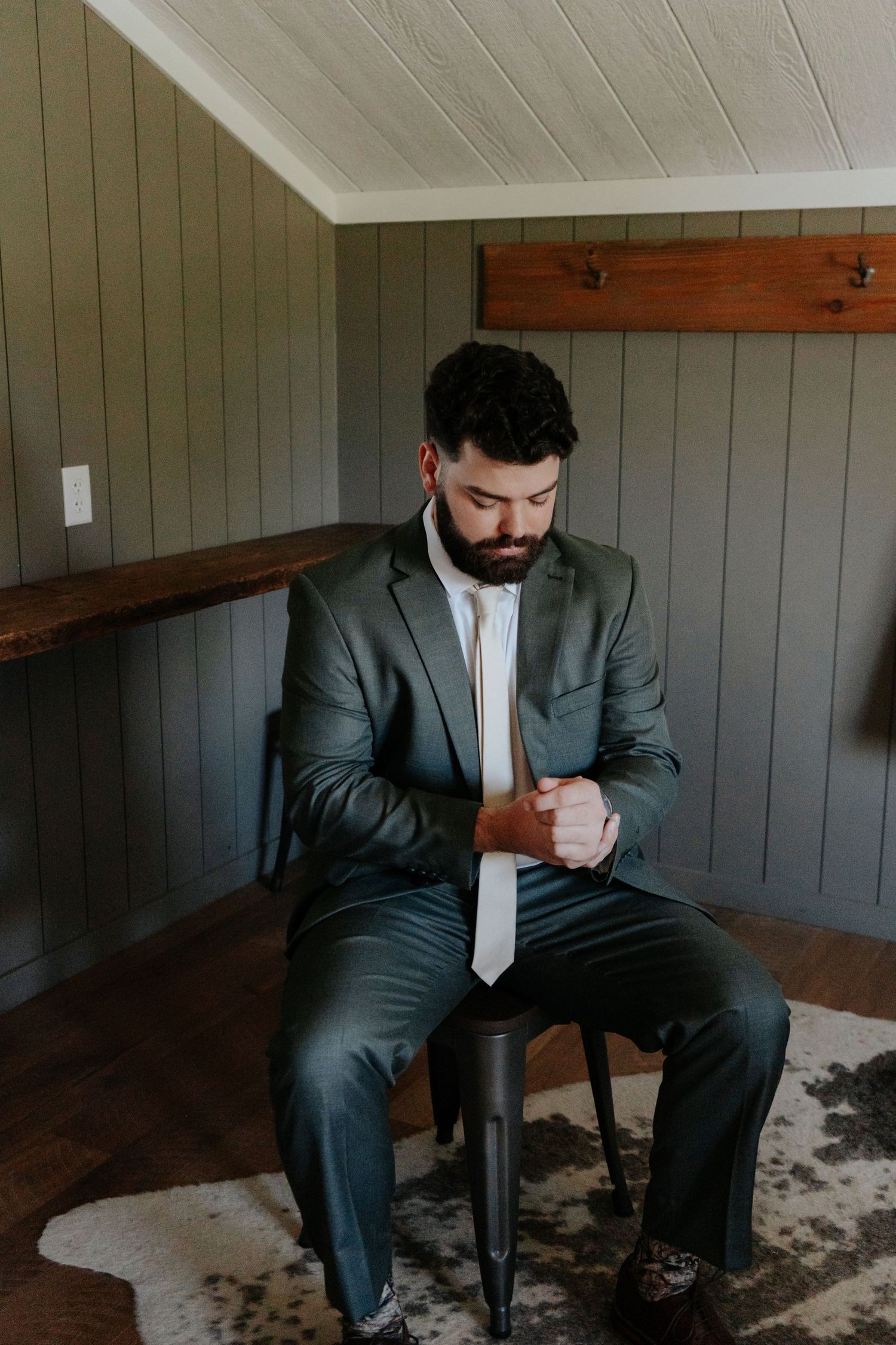 A bearded groom in a gray suit adjusts his cufflinks while seated on a stool in a rustic wood-paneled room.