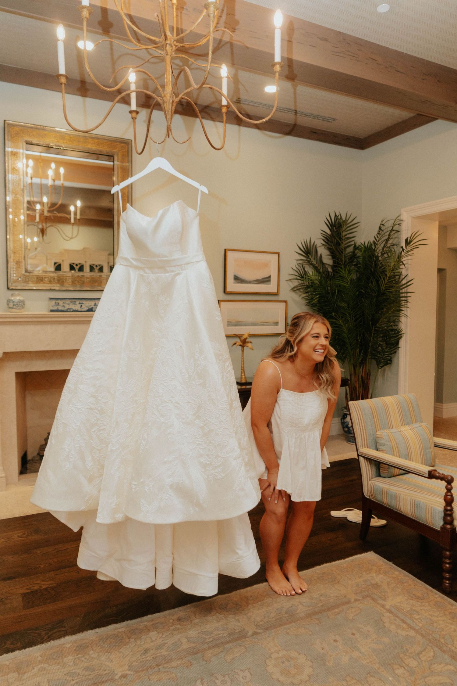 Bride in white romper smiling at her elegant strapless ball gown wedding dress hanging in a chandelier-lit room.