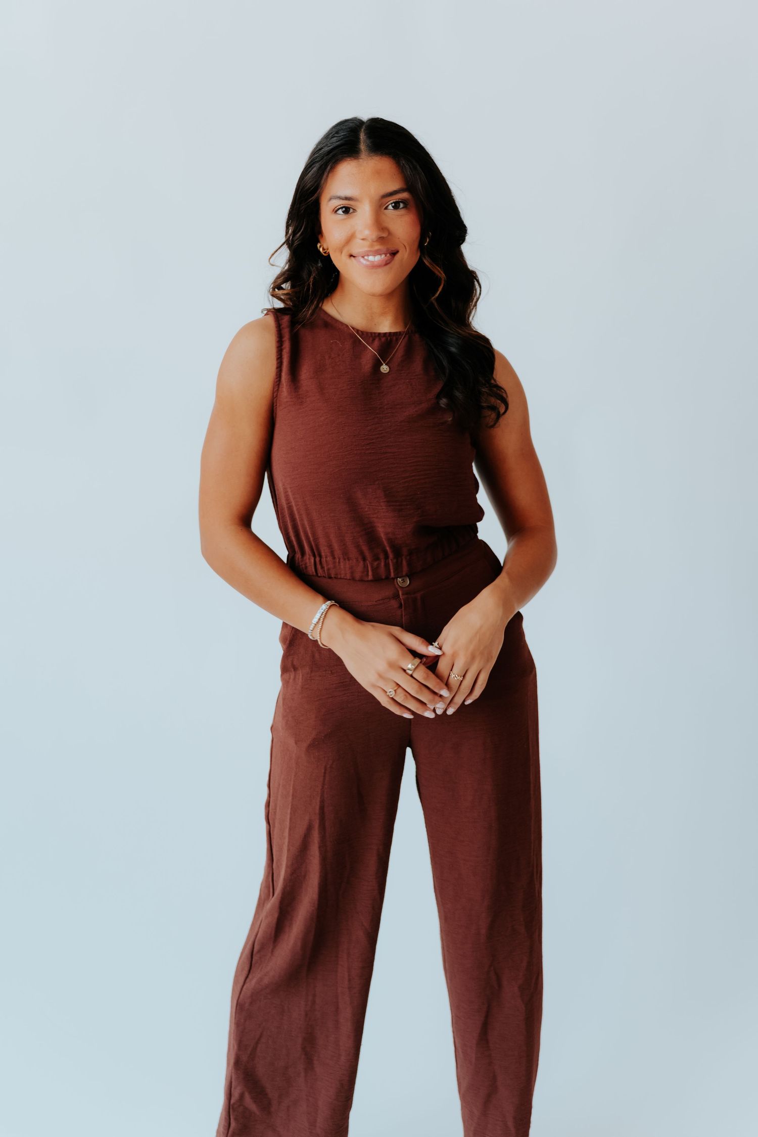 Woman wearing a brown sleeveless jumpsuit with wide-leg pants, smiling against a light gray background.
