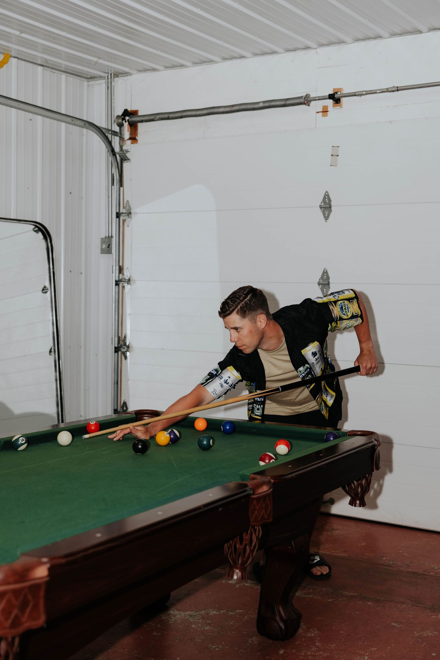 Man leaning over a pool table in a garage, lining up a shot with a cue stick on a green felt billiard table.
