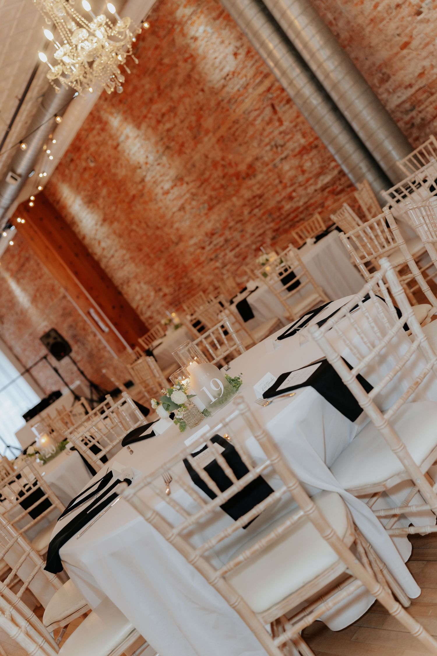 Elegant event venue with white tablecloths, black accents, chiavari chairs, chandelier, and exposed brick walls.