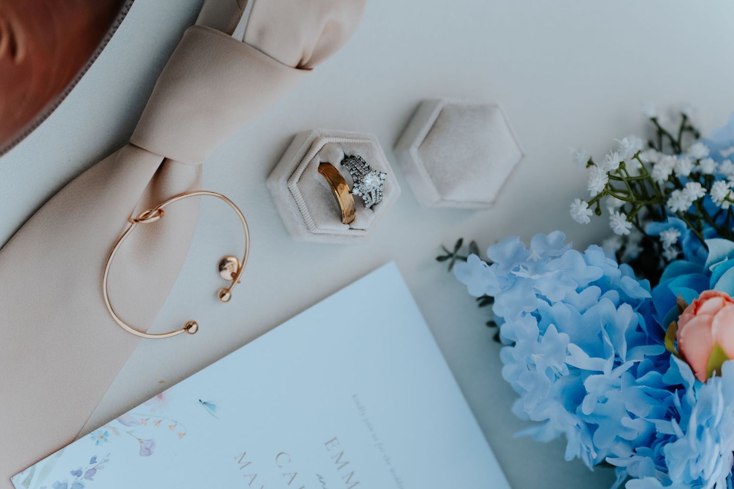 Elegant wedding flat lay with diamond ring in hexagon box, gold bracelet, blue hydrangeas, and open journal on white surface.