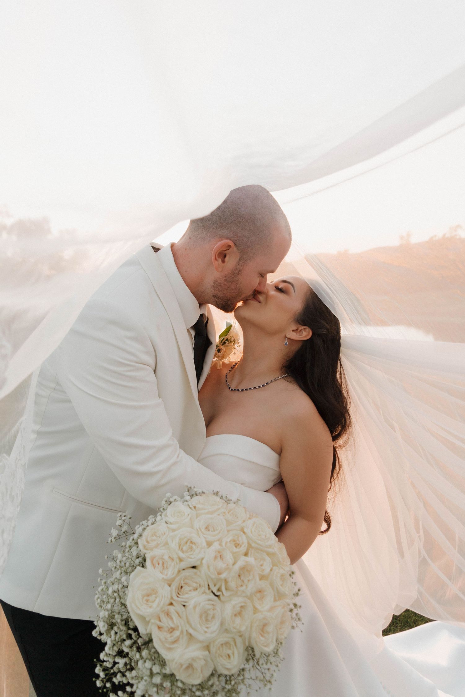 Bride in white gown and groom in white suit share romantic kisses with flowing veil and white rose bouquet.