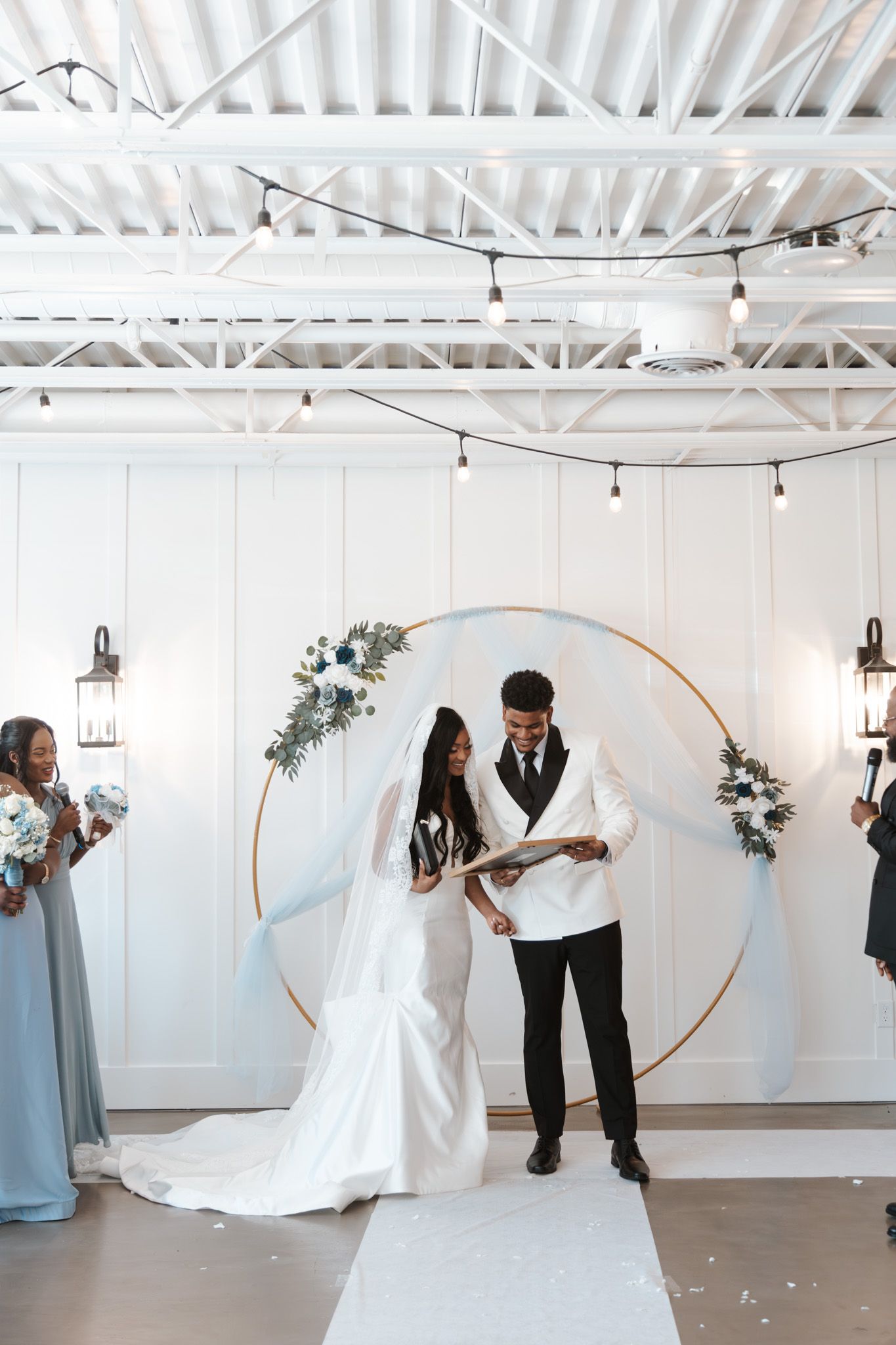 Bride and groom exchange vows under gold hoop arch with white shiplap backdrop at modern indoor wedding.