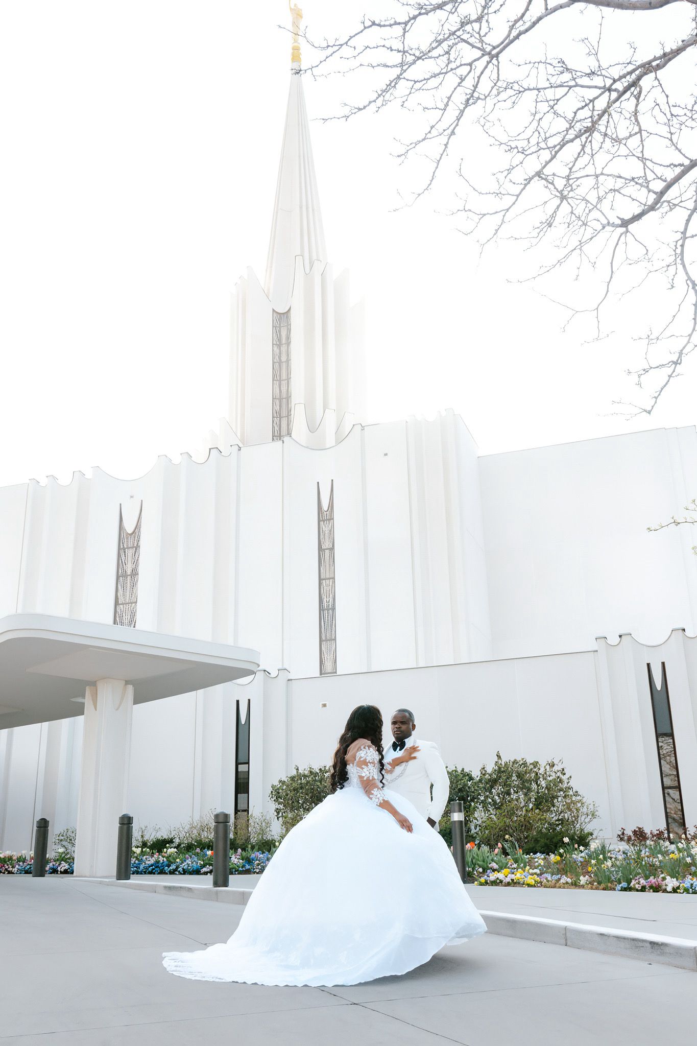 Bride and groom embrace outside a white LDS temple with a tall spire on their wedding day.