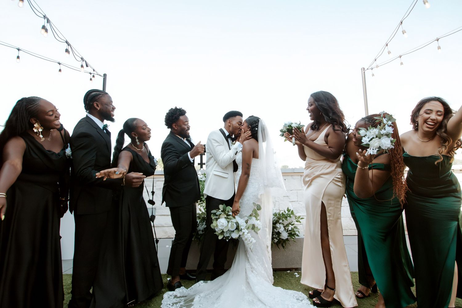 Wedding party in black and green gowns surrounds kissing bride and groom outdoors with string lights.