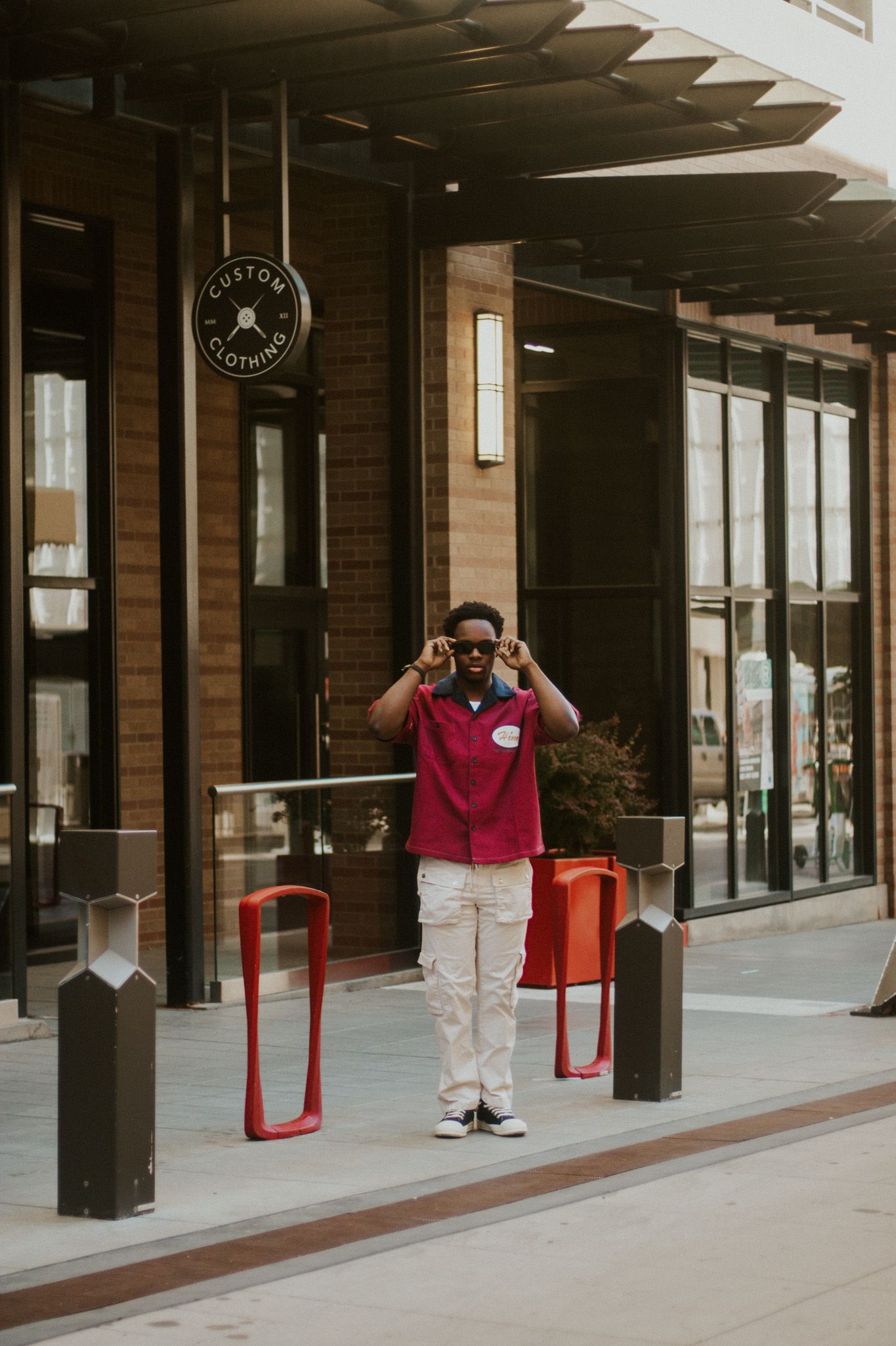 Young Black man in red shirt adjusting sunglasses while standing outside a custom clothing store.