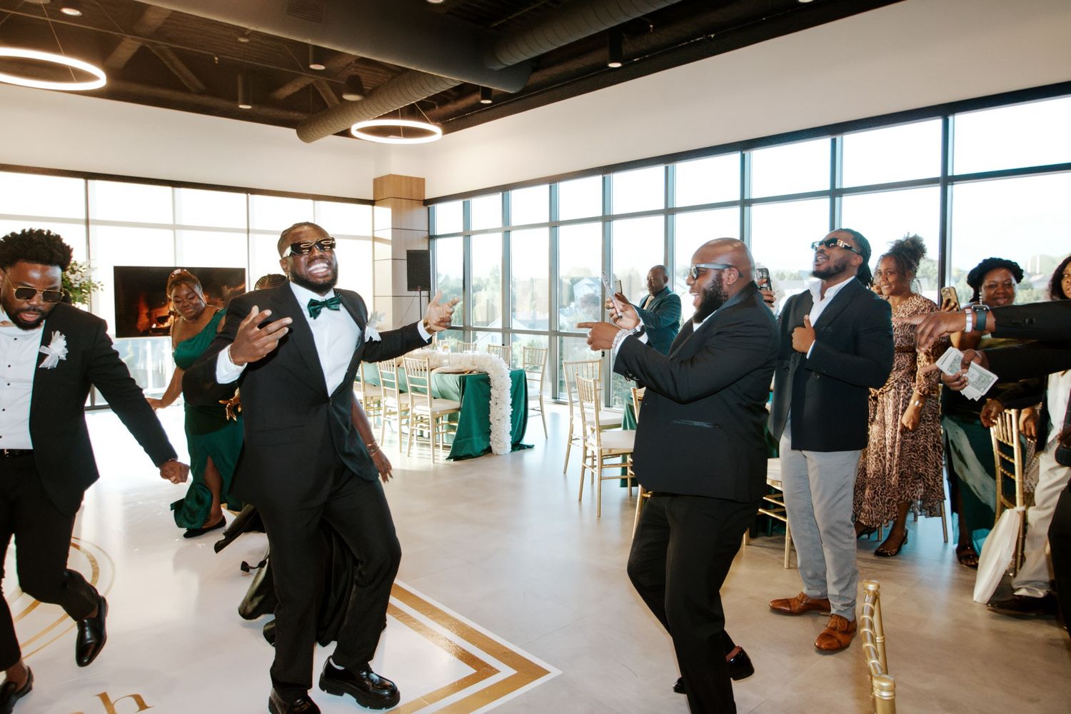 Groomsmen in black tuxedos dance energetically in modern venue with panoramic windows.
