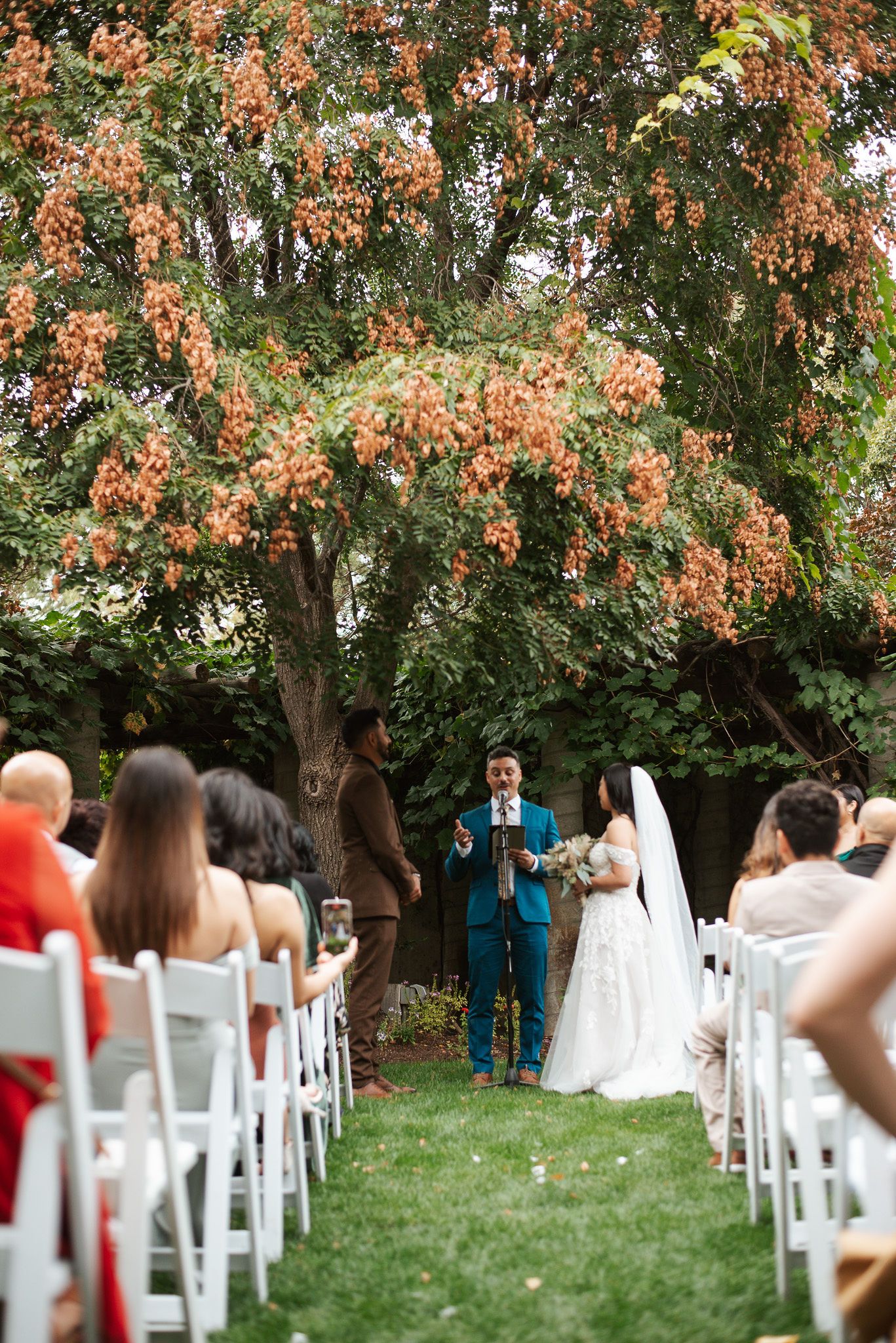 Father walks bride down aisle at outdoor garden wedding ceremony under autumn trees.