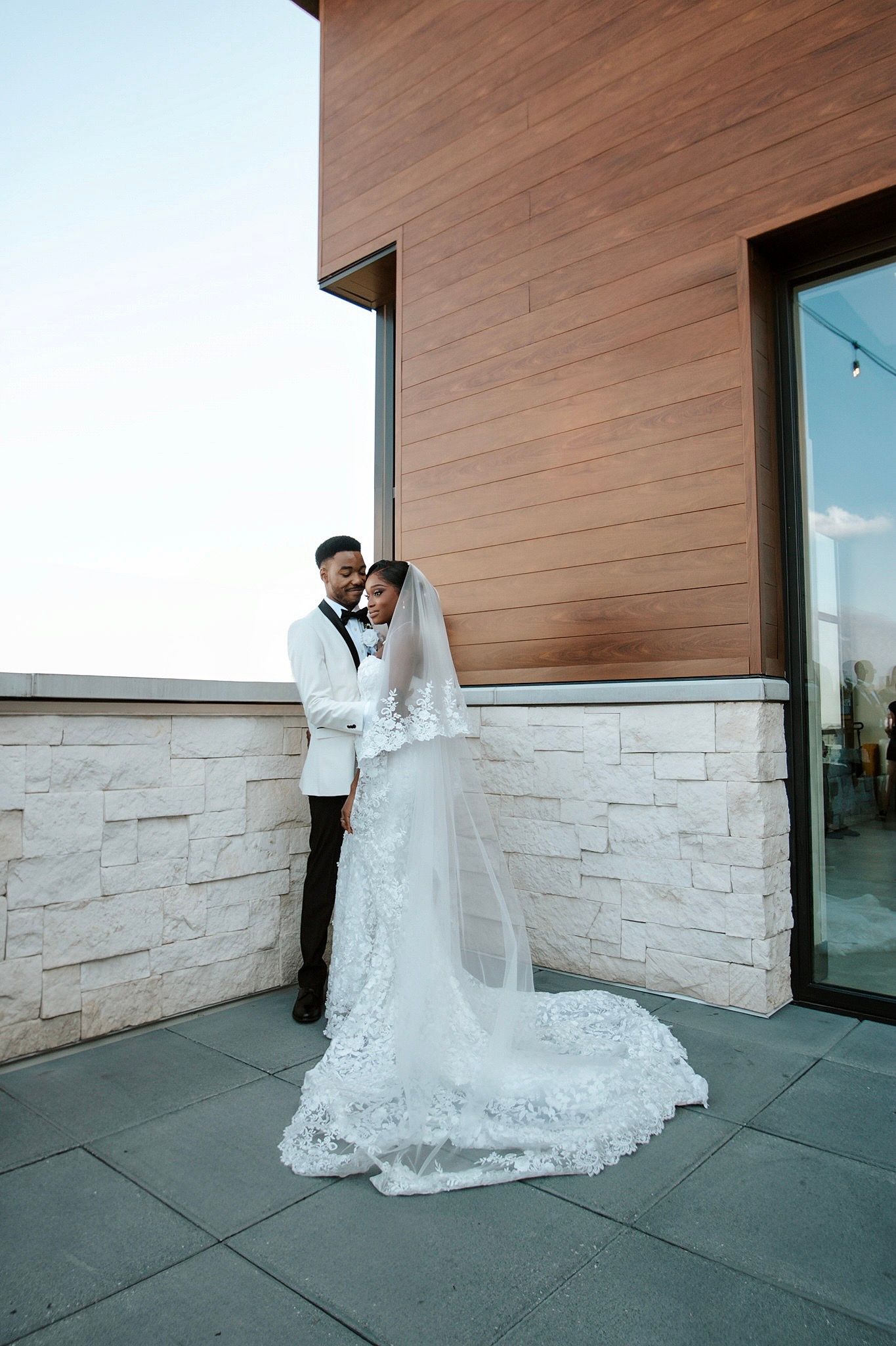 Bride in lace cathedral veil gown and groom in white tuxedo embrace on rooftop terrace at dusk.