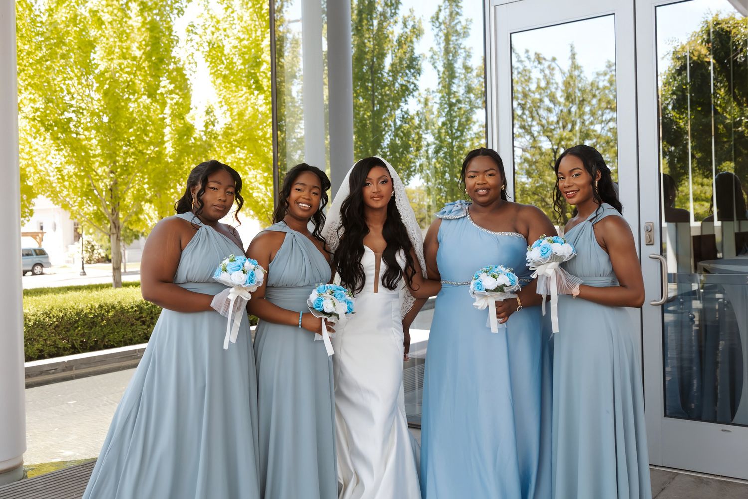 Bride in white gown poses with four bridesmaids in light blue dresses holding teal bouquets outdoors.