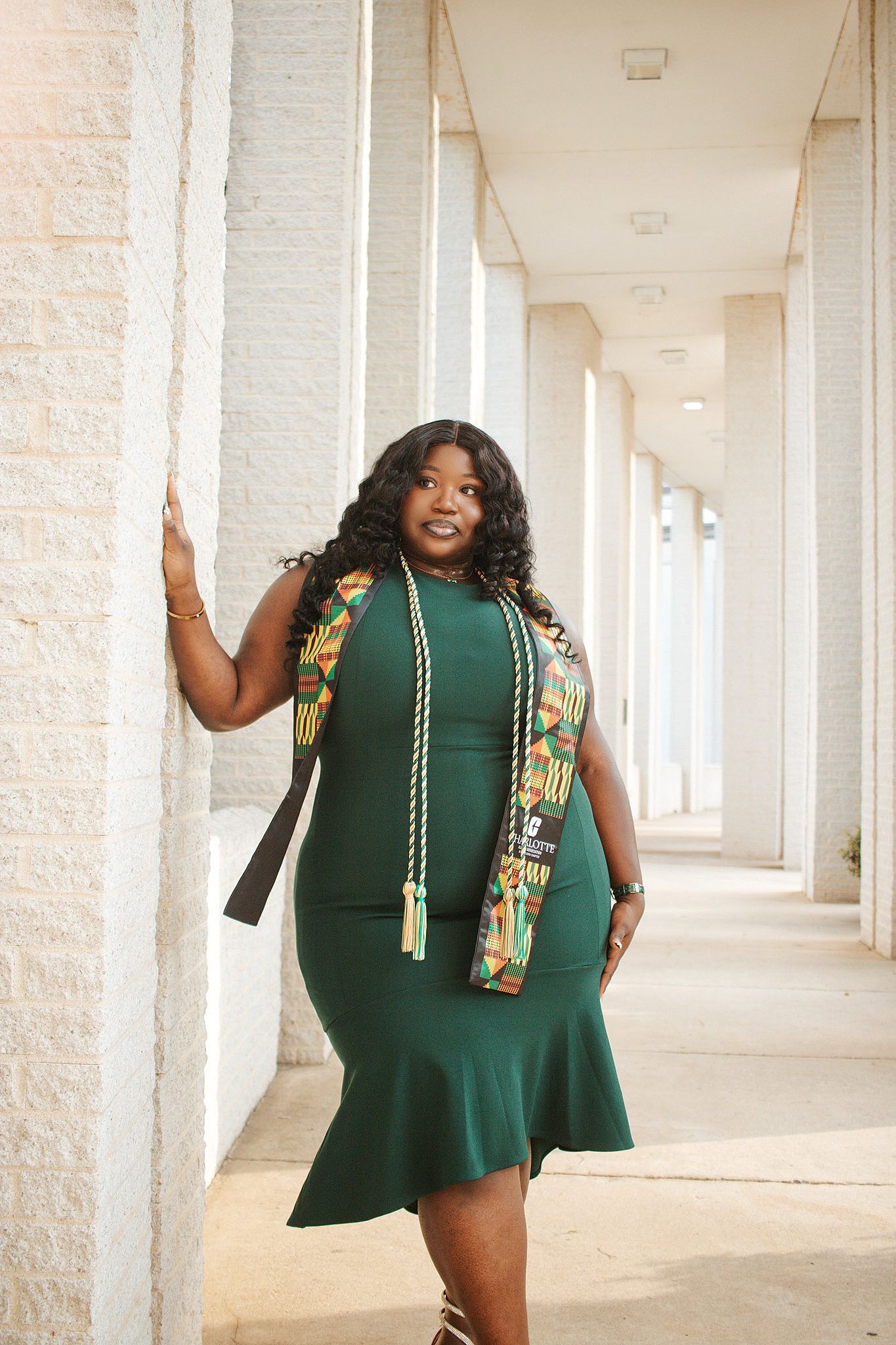 Black woman in green graduation dress with kente stole and honor cords posing in columned hallway.