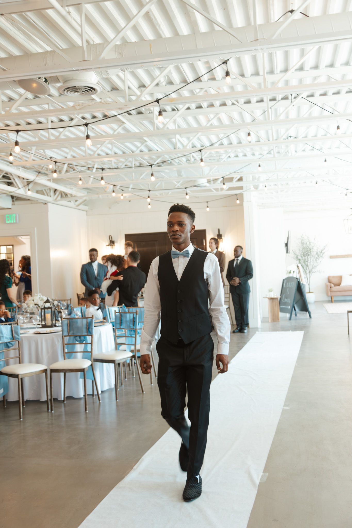 A groomsman in a black vest and bow tie walks down the aisle at an elegant wedding reception venue.