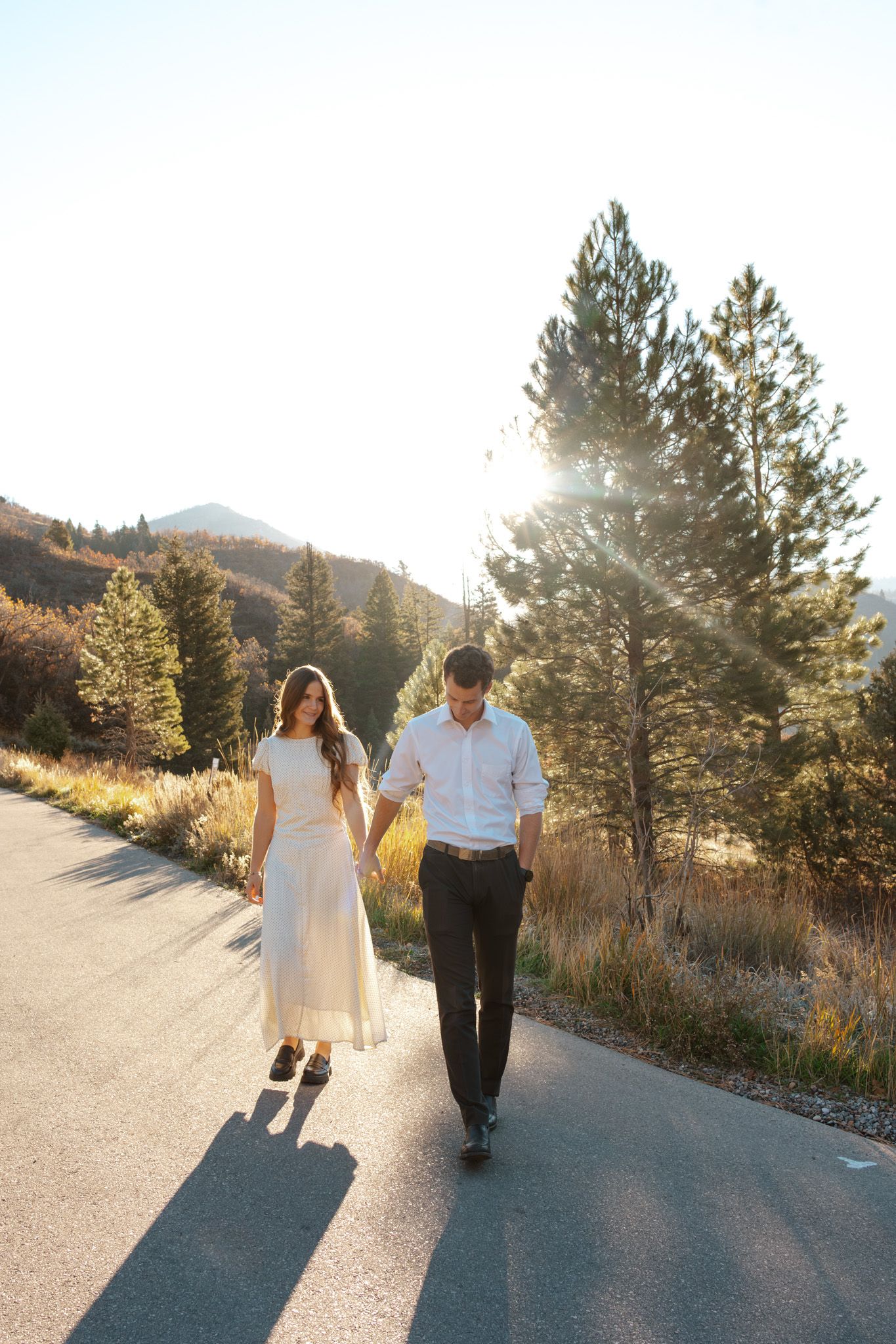 A couple holds hands walking down a sunlit mountain road surrounded by pine trees and golden light.