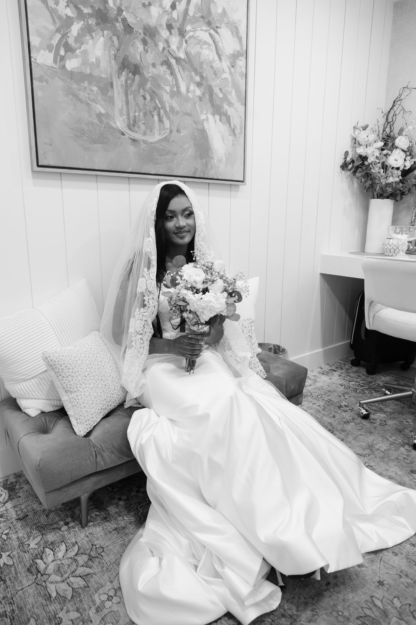 Bride in white gown and lace veil sits holding floral bouquet in elegant room, black and white photo.