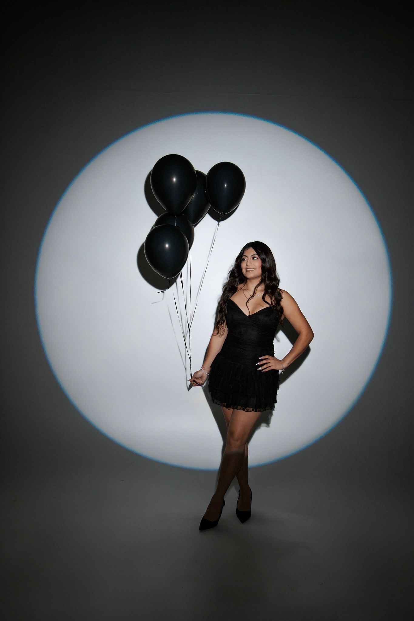 Woman in black mini dress holding black balloons in front of a circular studio light.
