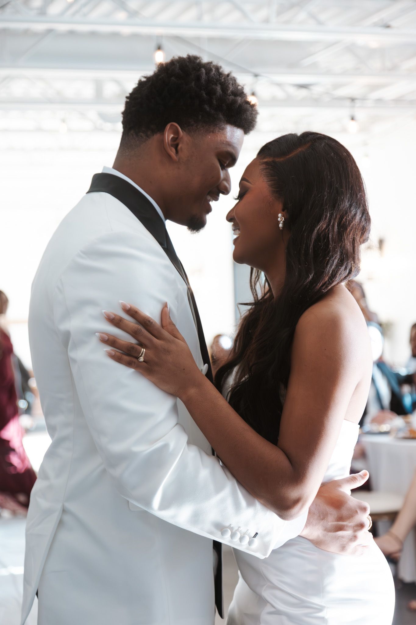 Newlywed couple shares romantic first dance, bride in white gown embracing groom in white tuxedo jacket.