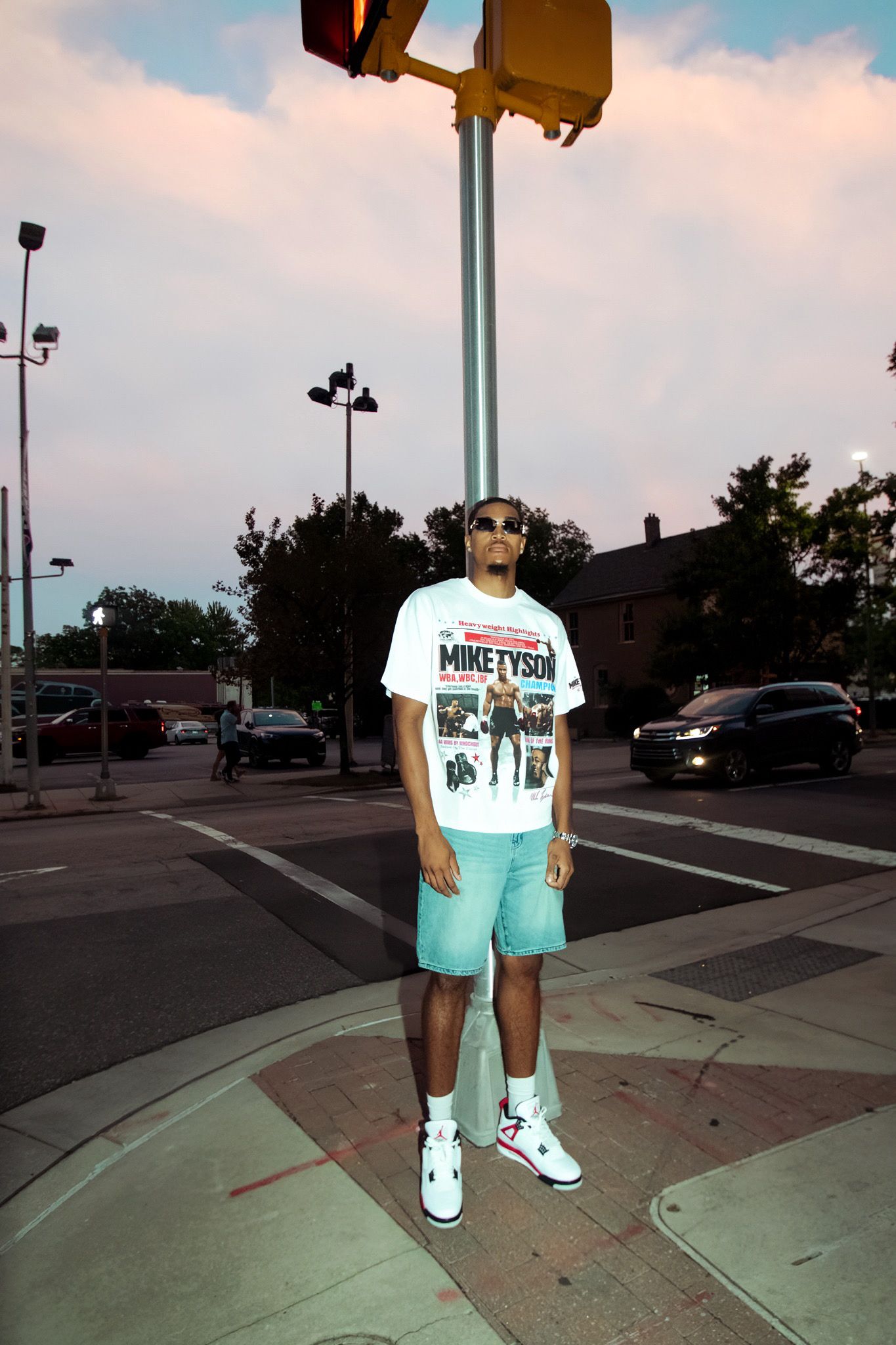 Young man in Mike Tyson graphic tee, mint shorts, and white Jordans standing at urban intersection.