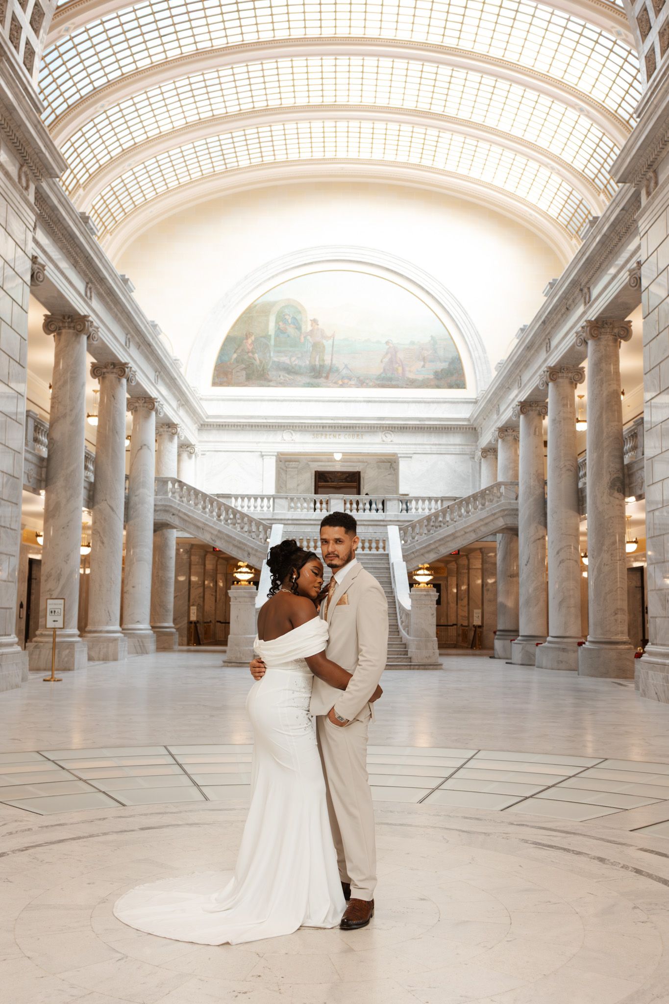 Interracial newlywed couple posing in grand marble rotunda with soaring columns and ornate painted arch.