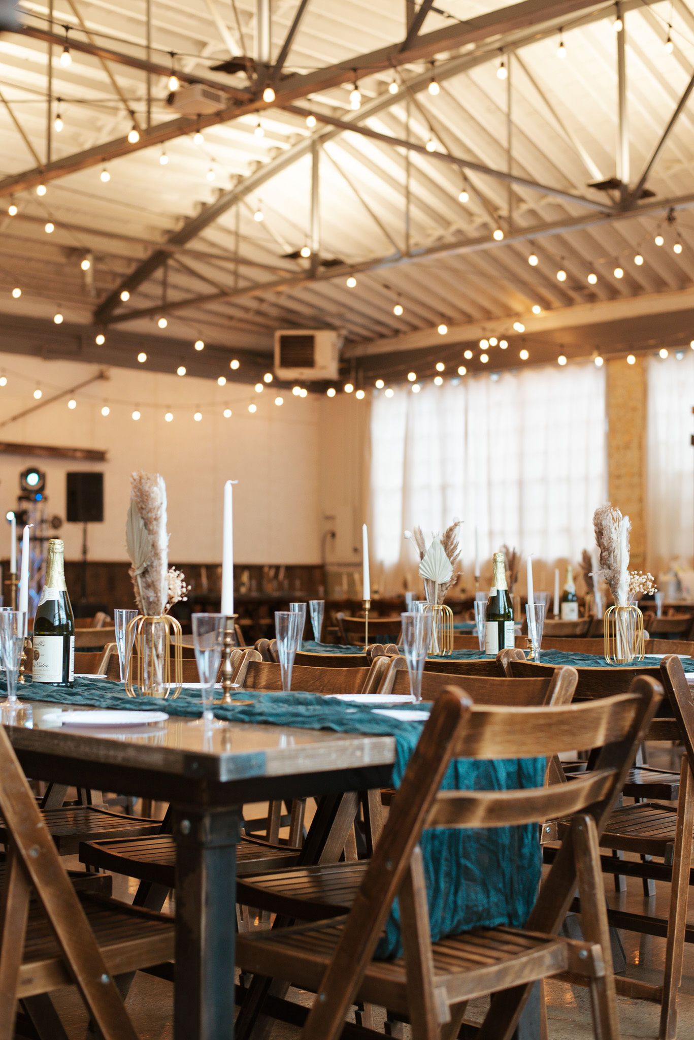 Rustic wedding reception hall with wooden tables, teal runners, candles, and string lights.