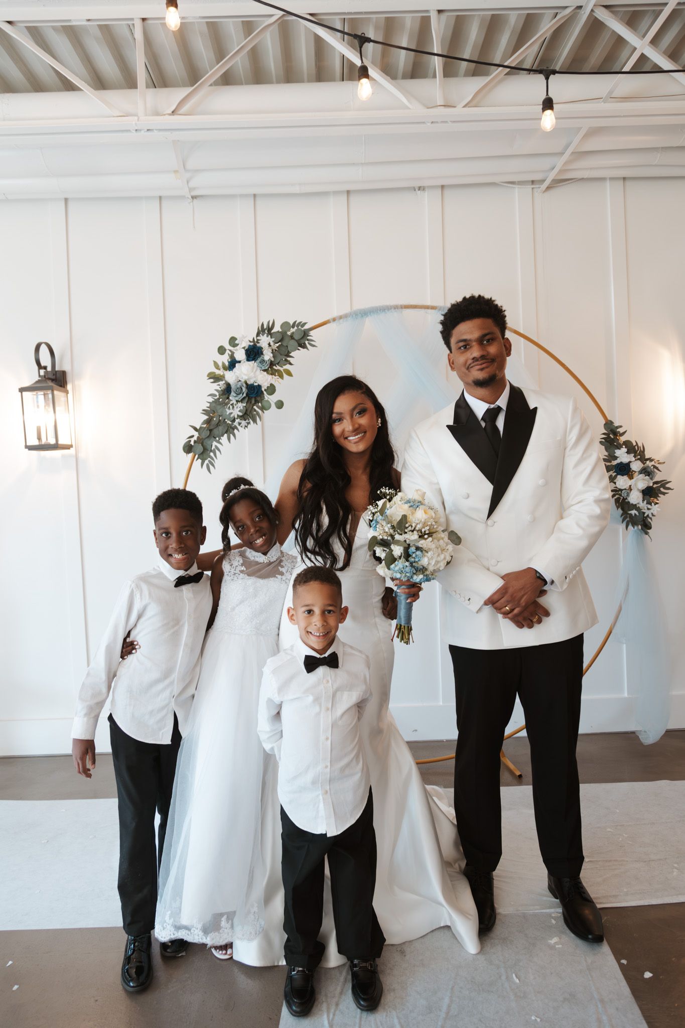 Bride and groom pose with three children in white attire before golden floral ceremony arch.