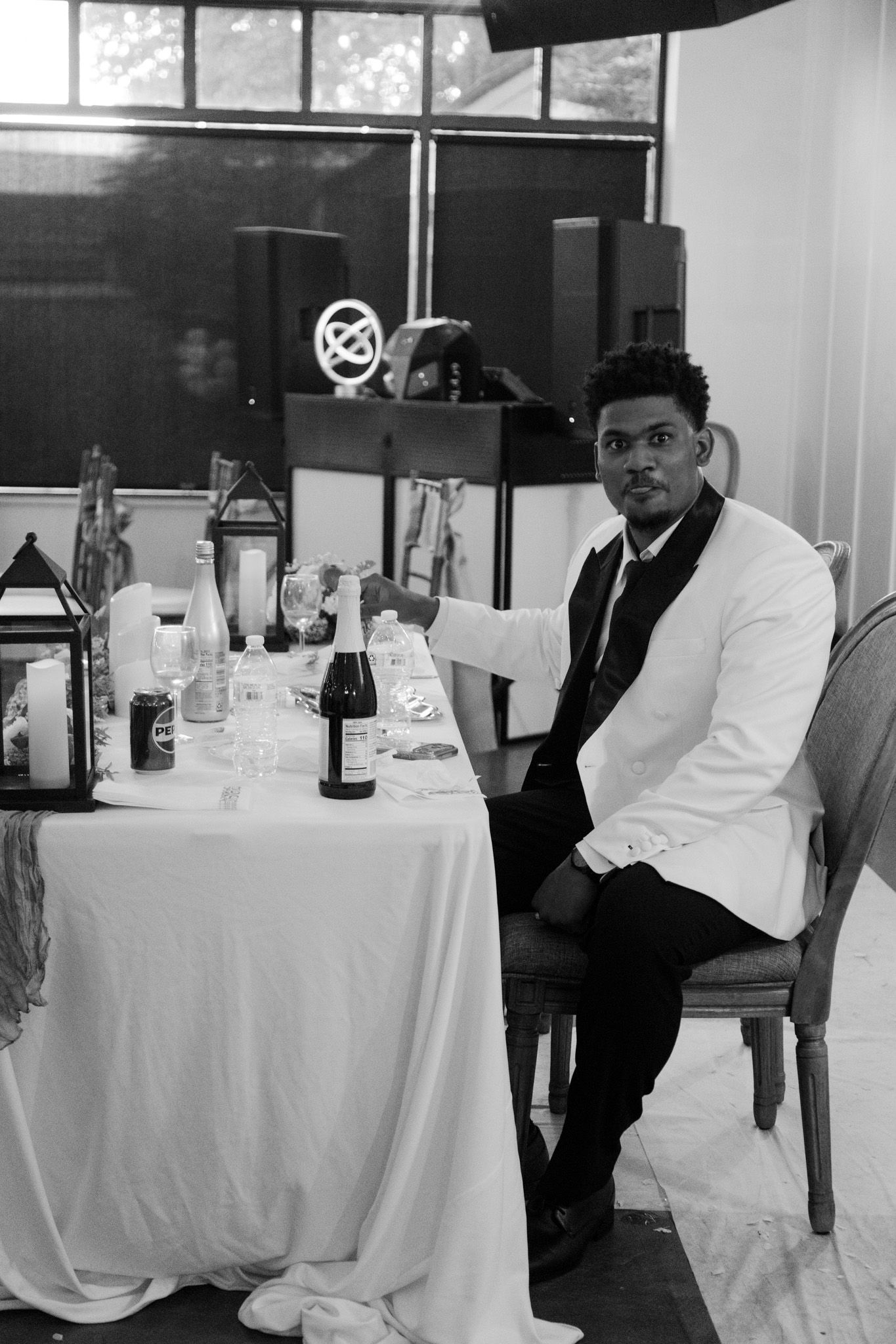 A groom in a white tuxedo jacket sits at a decorated reception table in this elegant black and white wedding photo.