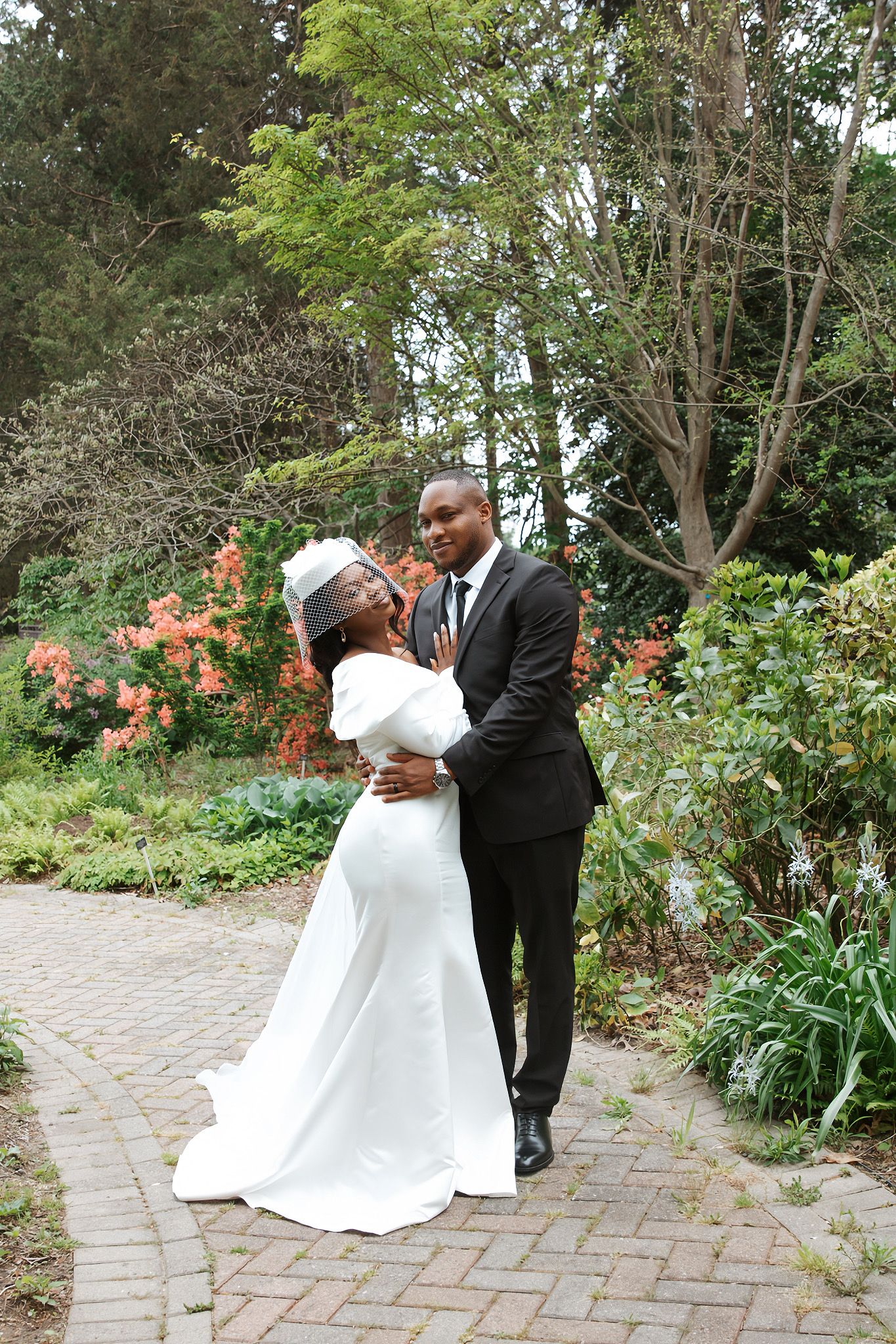 Bride in white off-shoulder gown and groom in black suit embrace on a garden brick pathway.