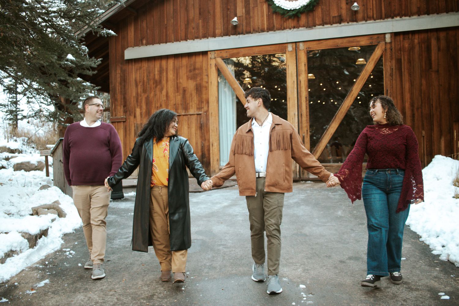 Family of four holding hands walking toward a rustic wooden barn in a snowy winter landscape.