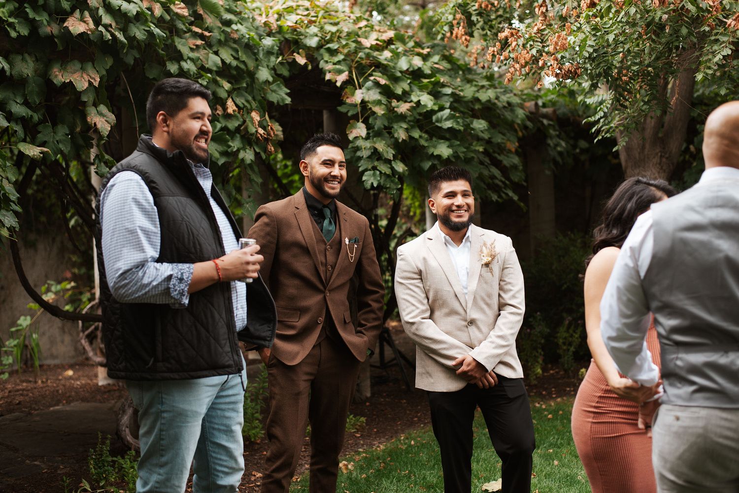 Three men in wedding attire smile and chat in lush ivy-covered outdoor garden setting.