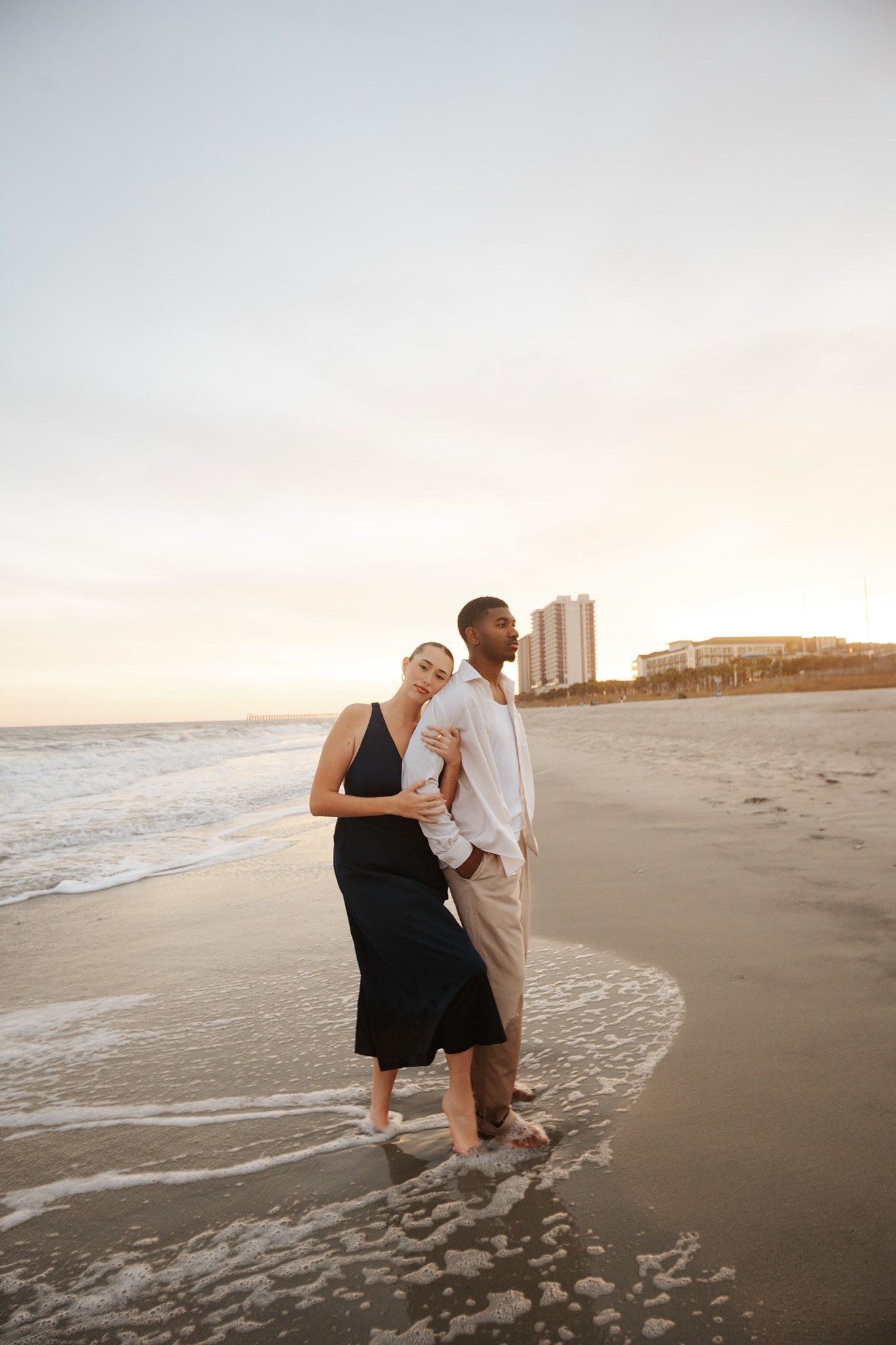 A couple embraces romantically on a sandy beach at sunset with ocean waves behind them.