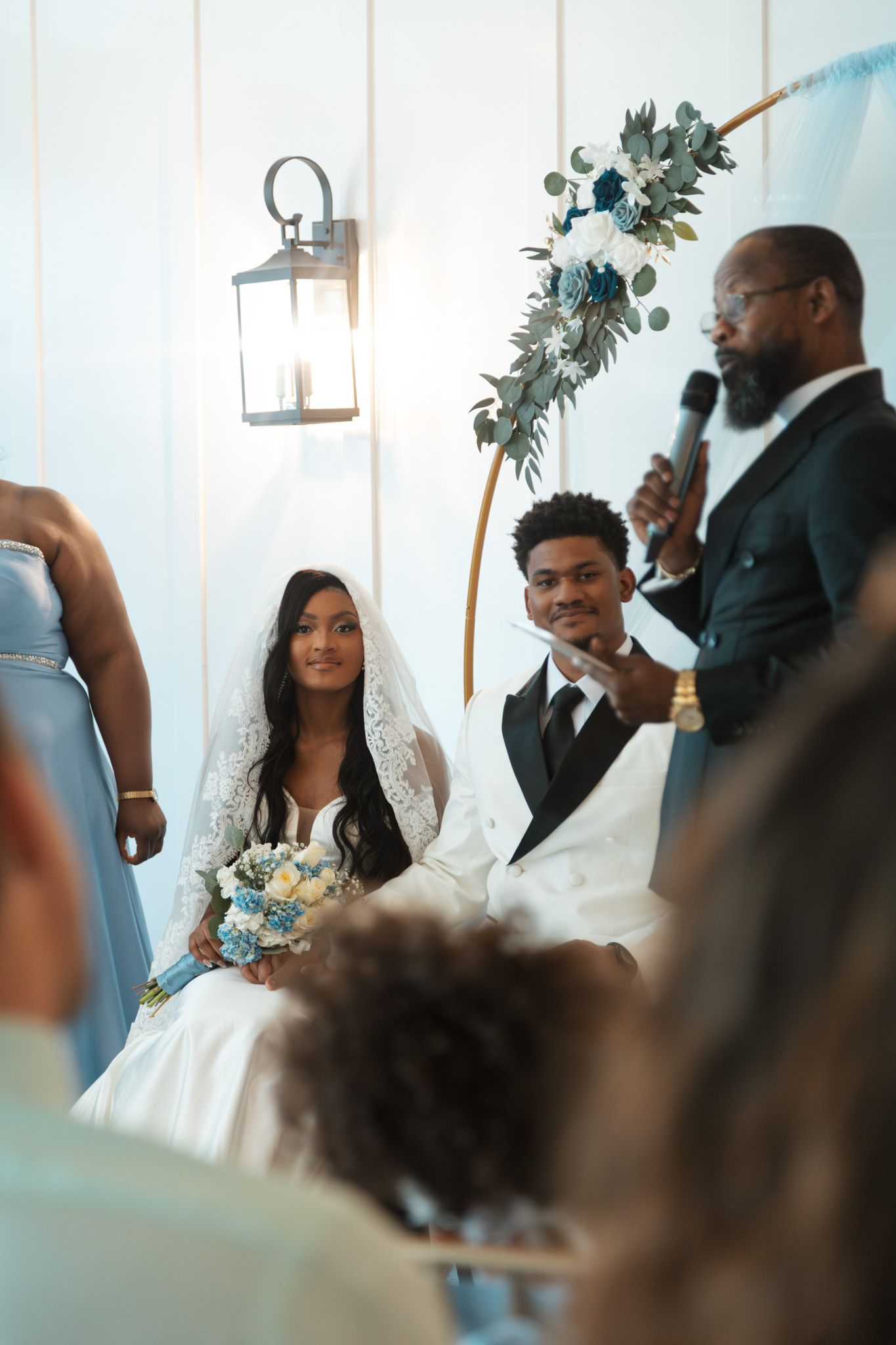 A bride and groom sit together during their wedding ceremony while an officiant speaks into a microphone beside a floral arch.