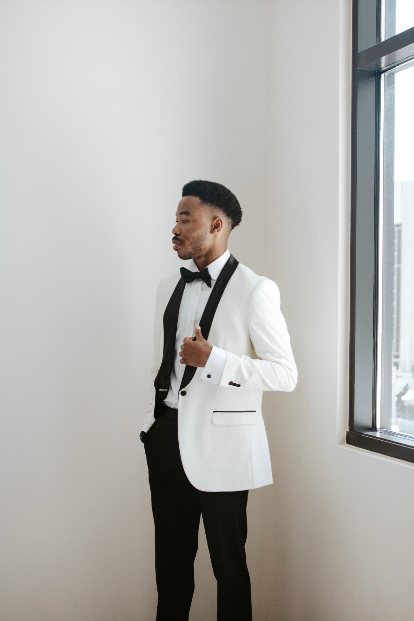 Dapper groom in white tuxedo jacket with black bow tie standing by window in elegant room.