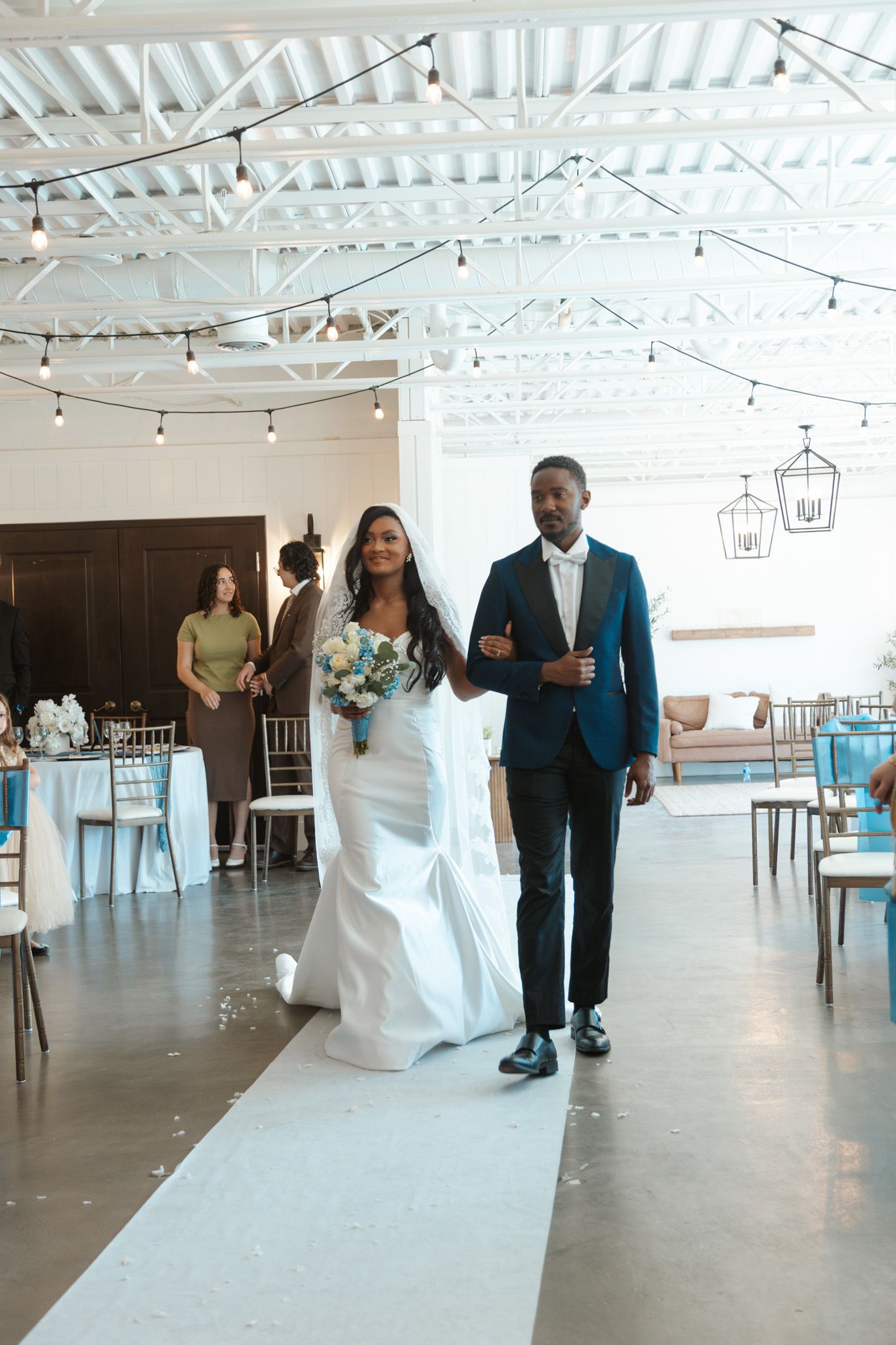 Bride in white gown walks down aisle with escort at elegant indoor wedding ceremony.
