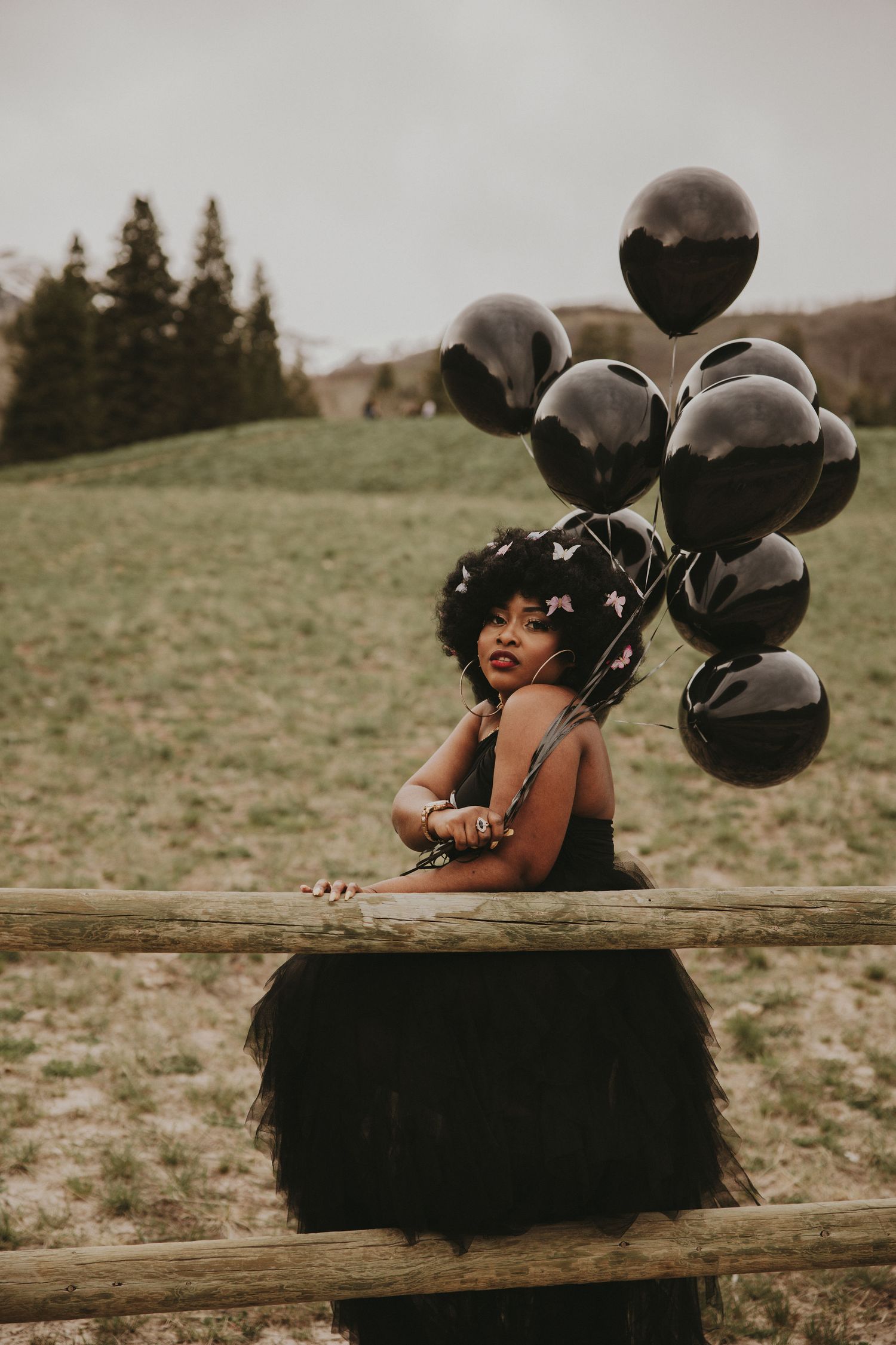 Black woman in black tutu skirt holding black balloons while leaning on rustic wooden fence outdoors.