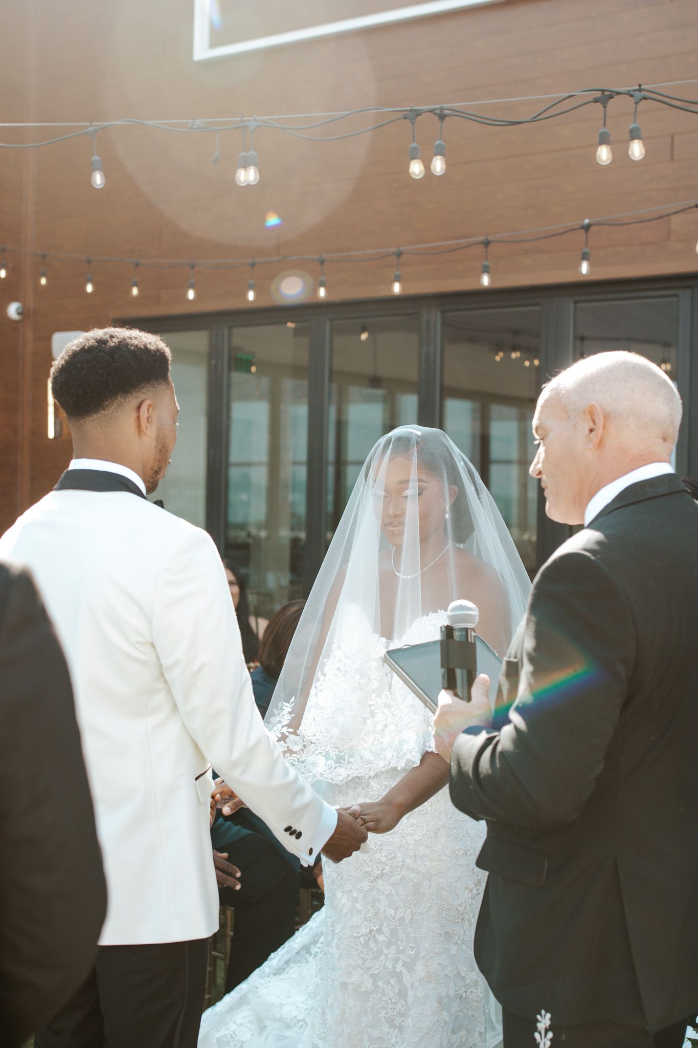 Bride and groom hold hands at outdoor wedding ceremony with string lights, officiant reading vows in sunlit setting.