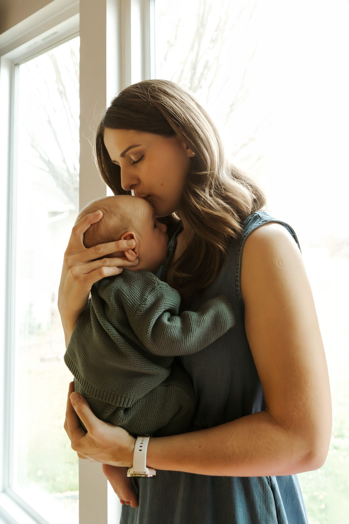 A mother tenderly kisses her newborn baby wrapped in a green sweater near a bright window.