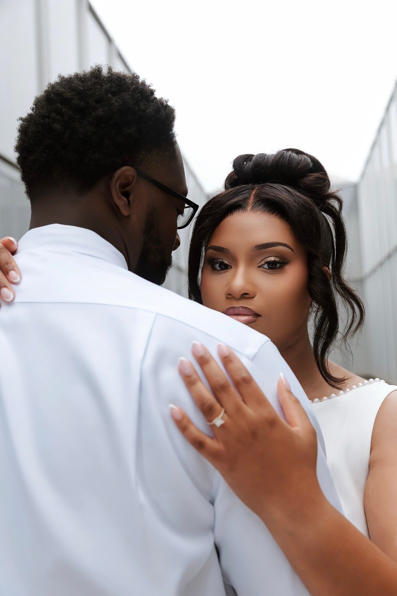 Black couple in white attire embrace outdoors, woman displaying diamond engagement ring while gazing at camera.
