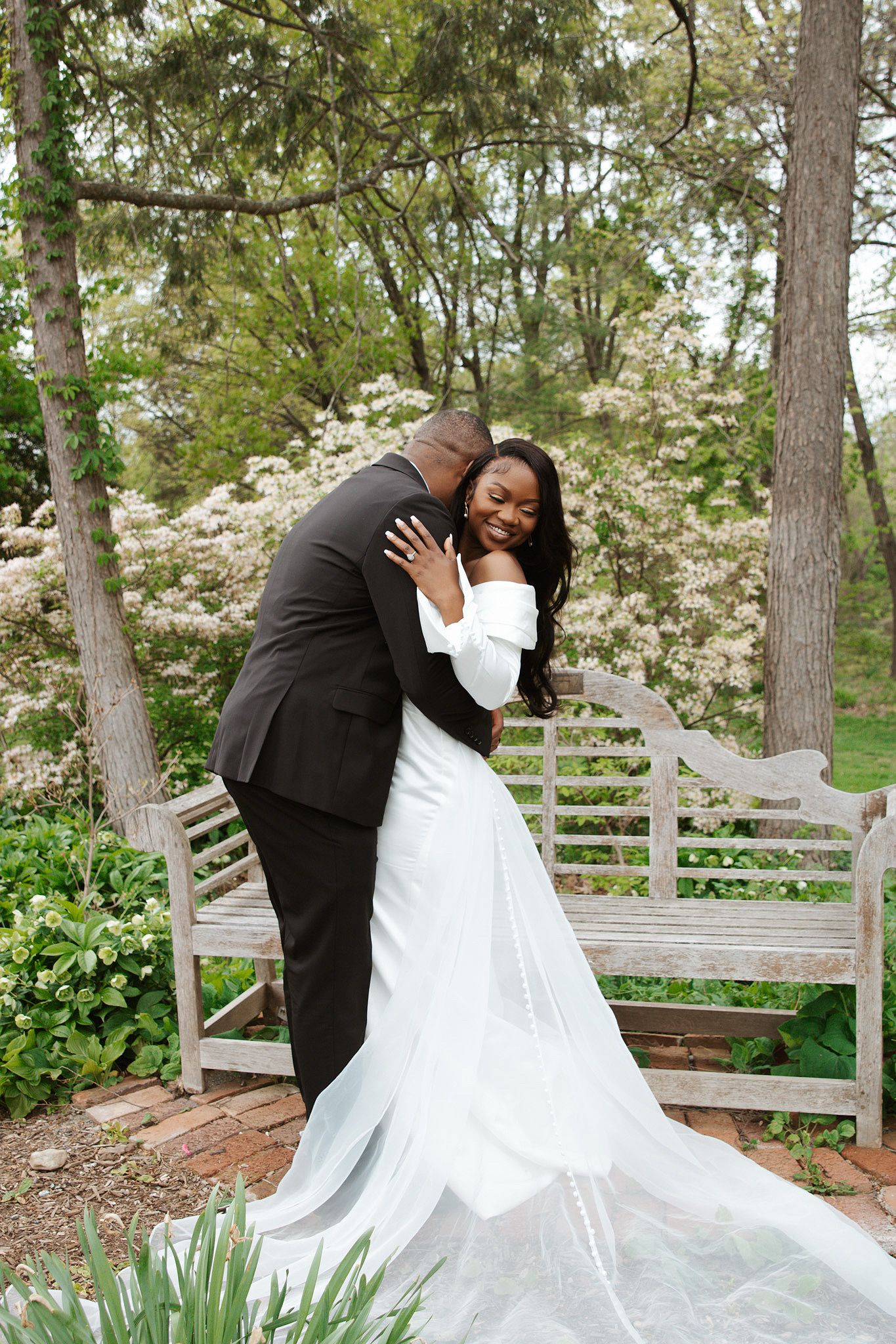 Black couple embraces in garden wedding photo, bride in white off-shoulder gown with veil, groom in black suit.
