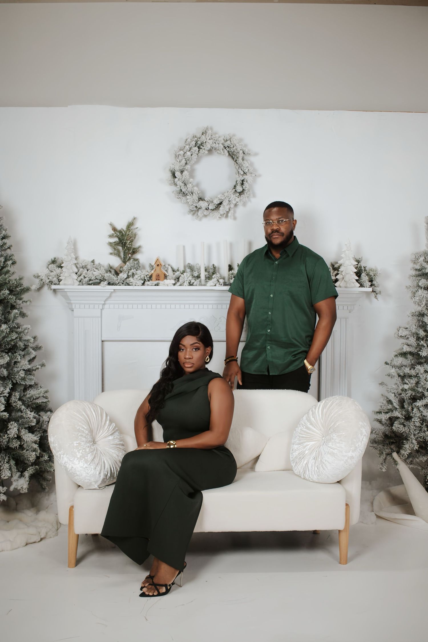 Couple posing in matching green outfits before a white fireplace with Christmas holiday decor.
