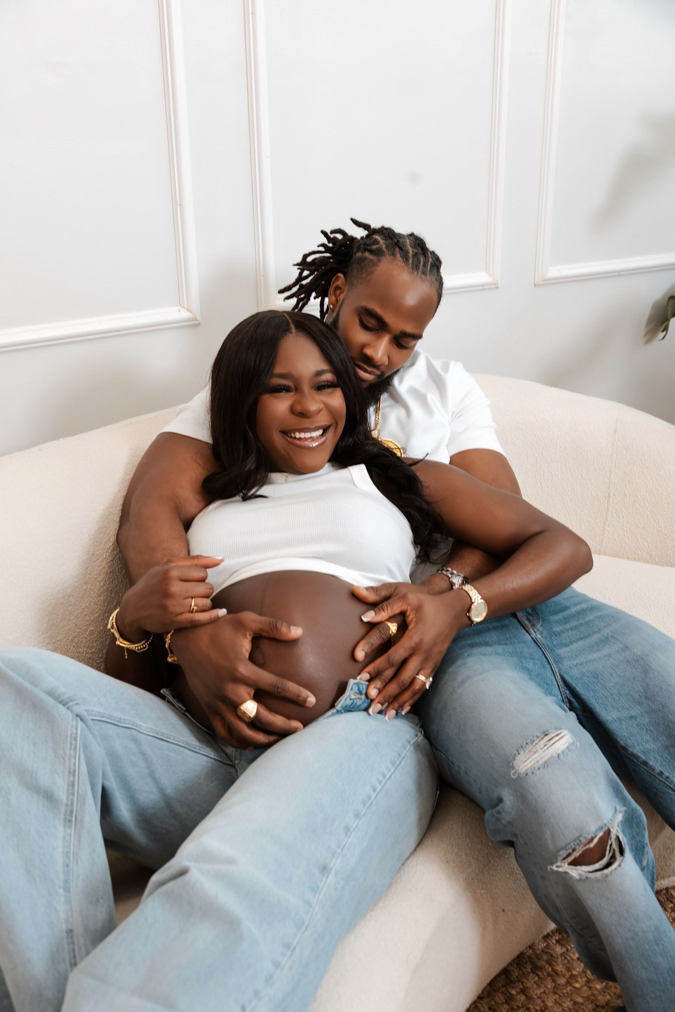 Happy pregnant couple sitting on sofa, man embracing woman from behind, both touching baby bump, wearing jeans.